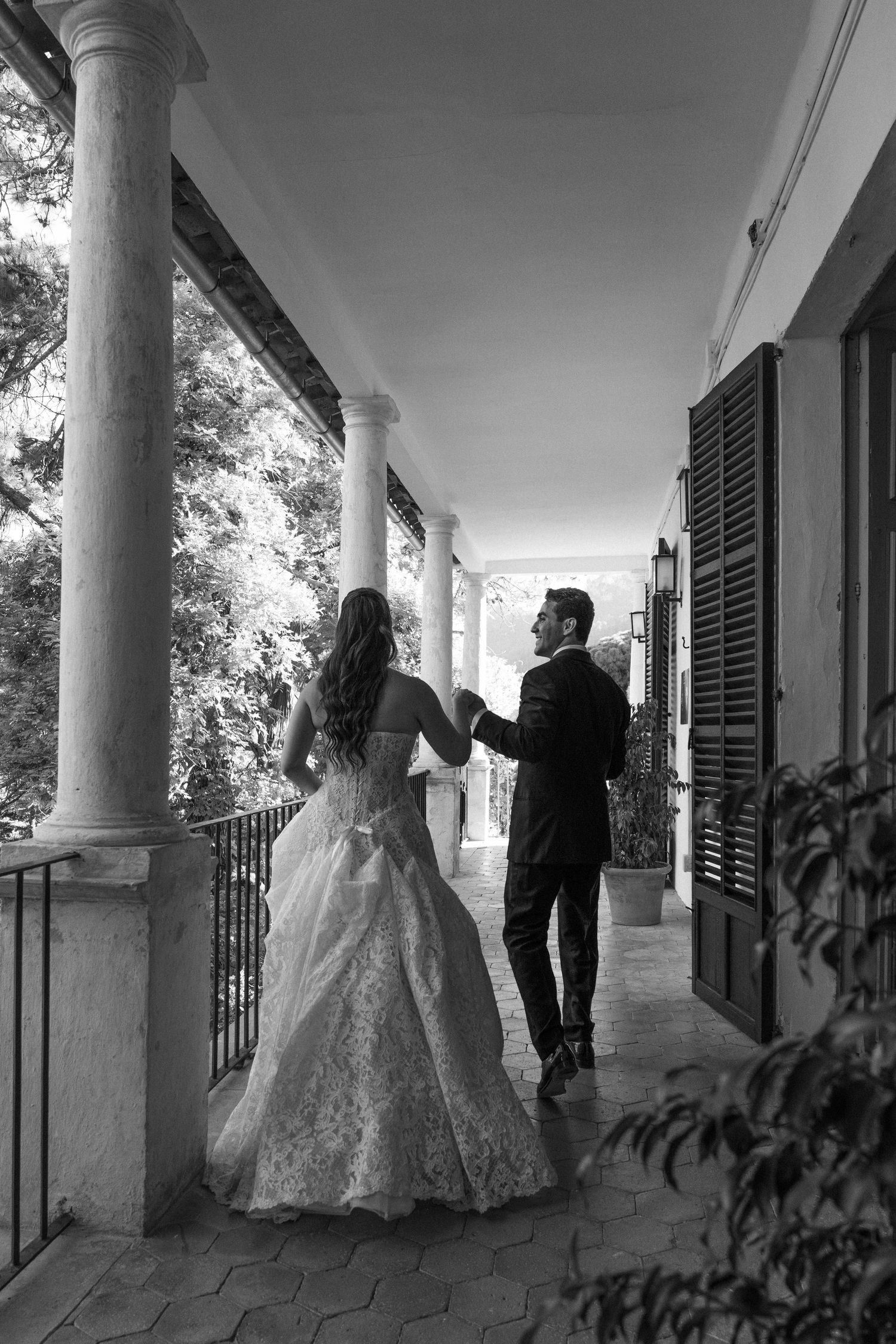 Black and white photo of a bride and groom walking hand in hand along an elegant columned veranda.