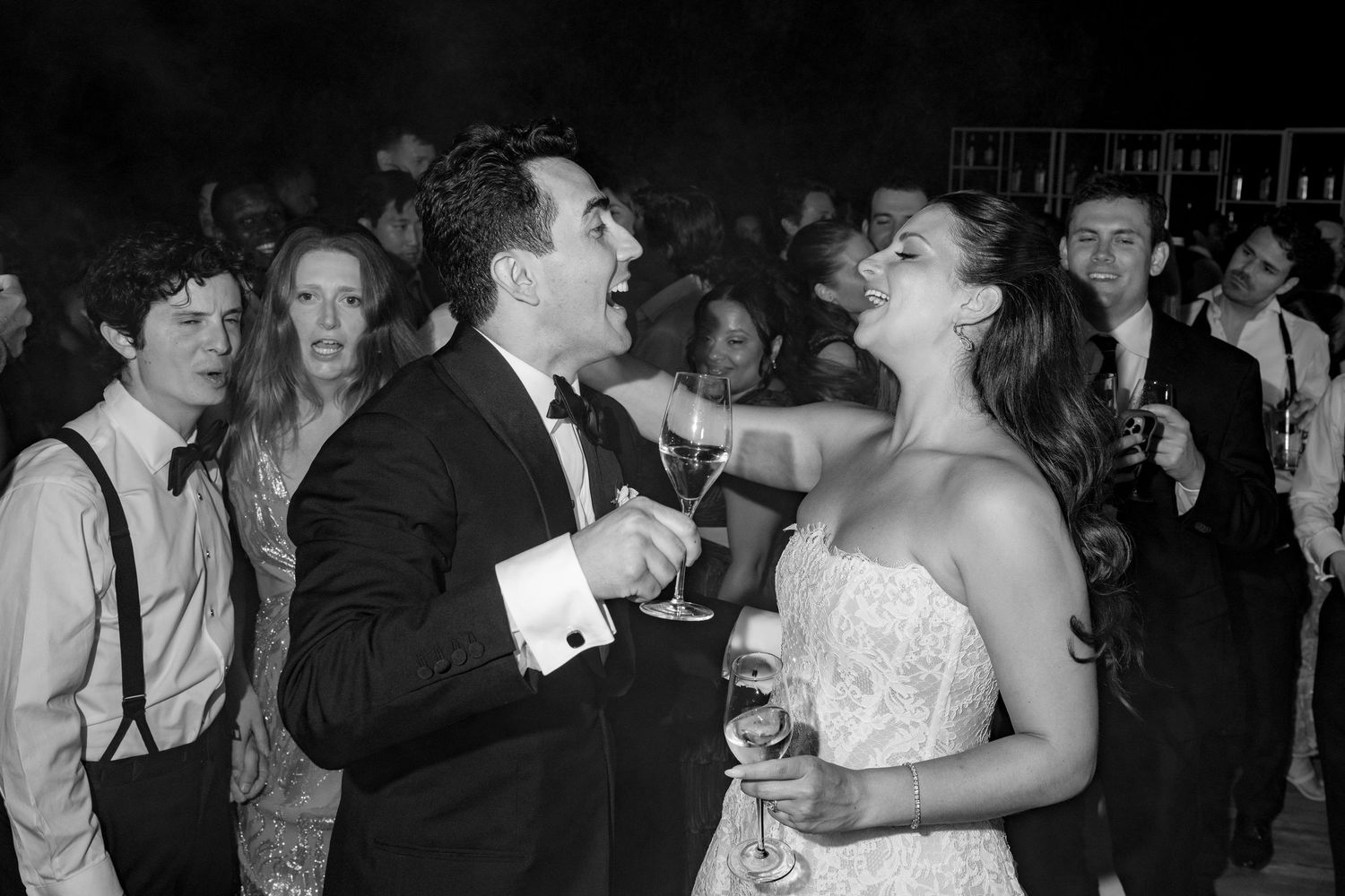 Black and white photo of elegantly dressed guests laughing and dancing at a lively wedding reception party.