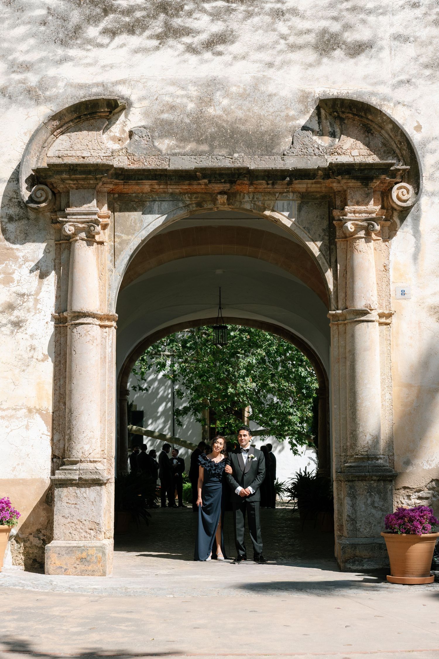 Couple in formal attire standing under ornate stone archway at historic European venue during wedding celebration.