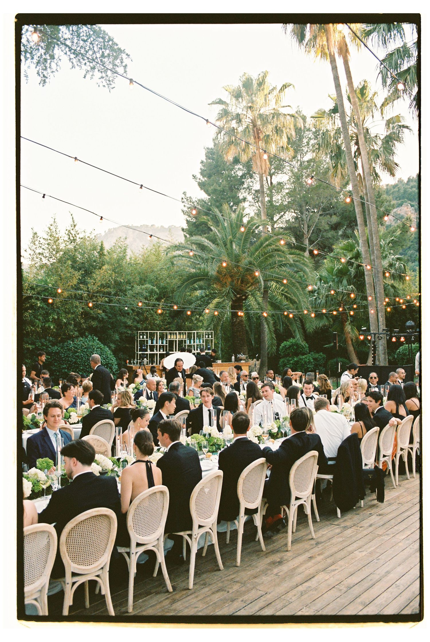 Elegant outdoor wedding reception on a wooden deck surrounded by palm trees, with string lights and guests seated at long tables.