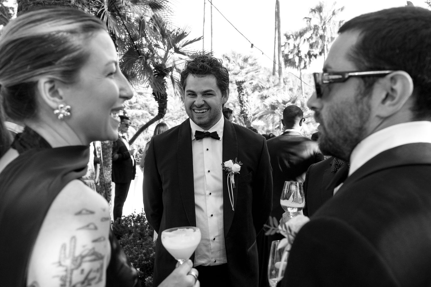 Groom in tuxedo smiling with guests at outdoor wedding reception in black and white photo.