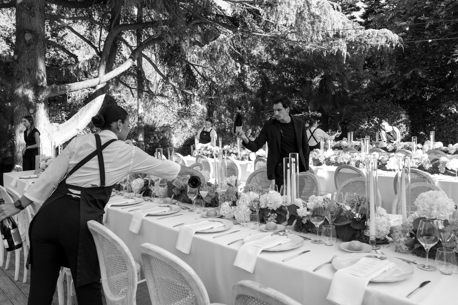 Black and white photo of elegantly set outdoor banquet tables with staff arranging floral centerpieces and glassware under trees.