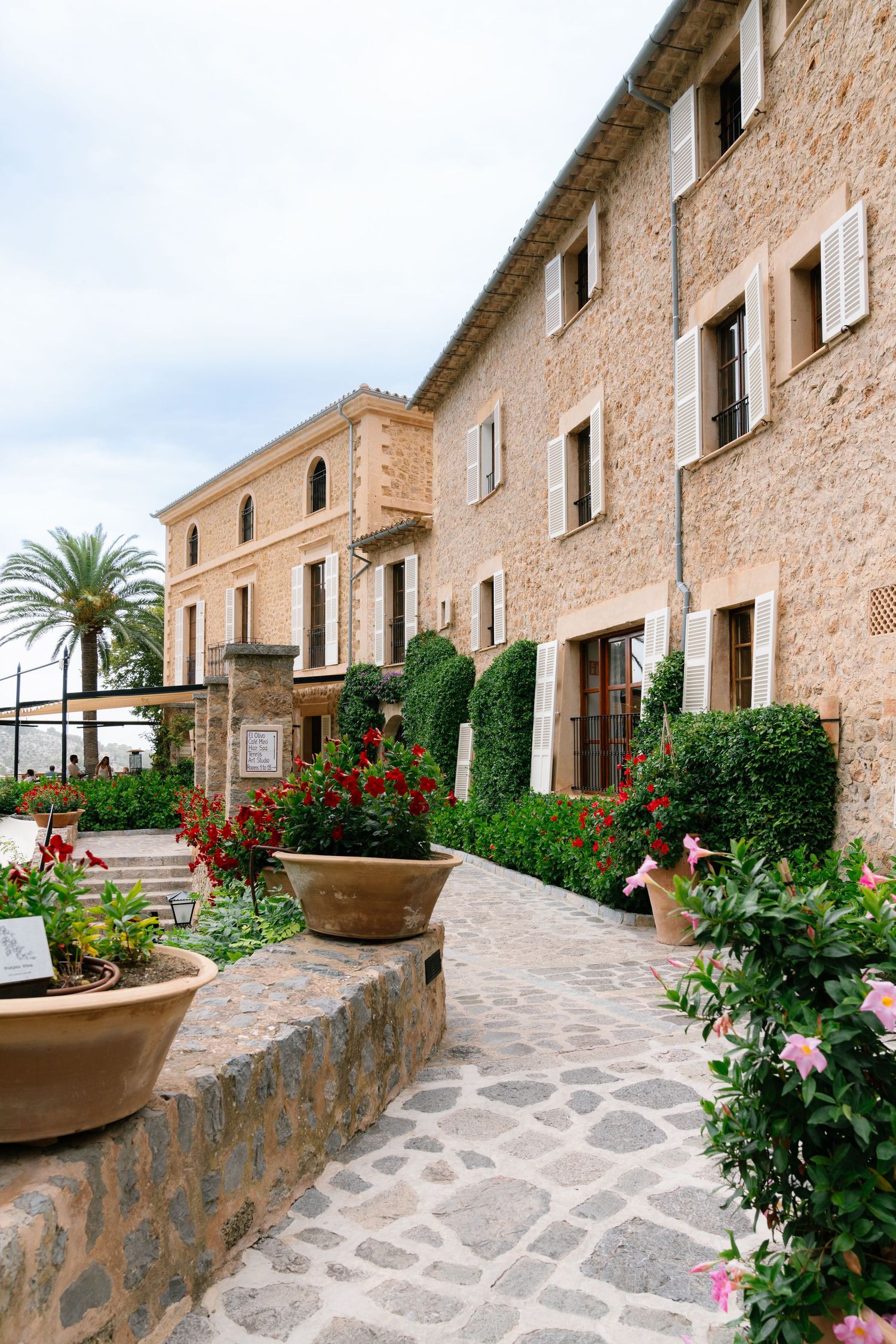 Stone building with lush garden pathway lined with red flowers and green shrubs under a cloudy sky at Belmond La Residencia.