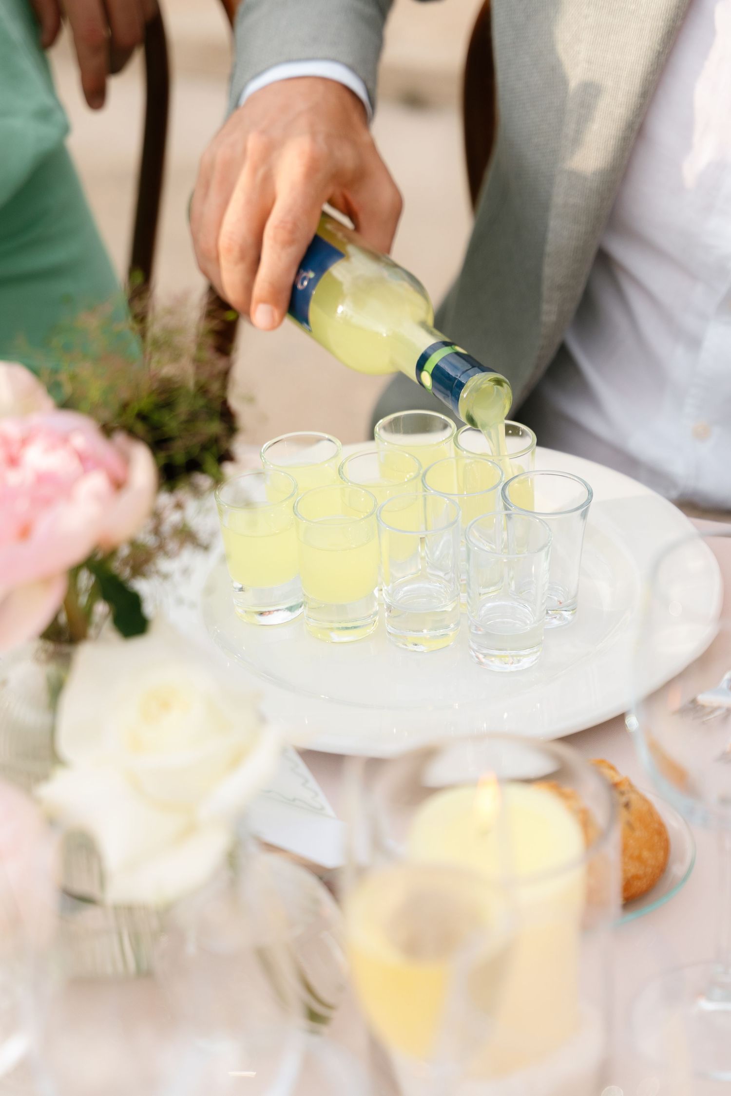 Hands pouring yellow cocktails into small glasses at an outdoor event.