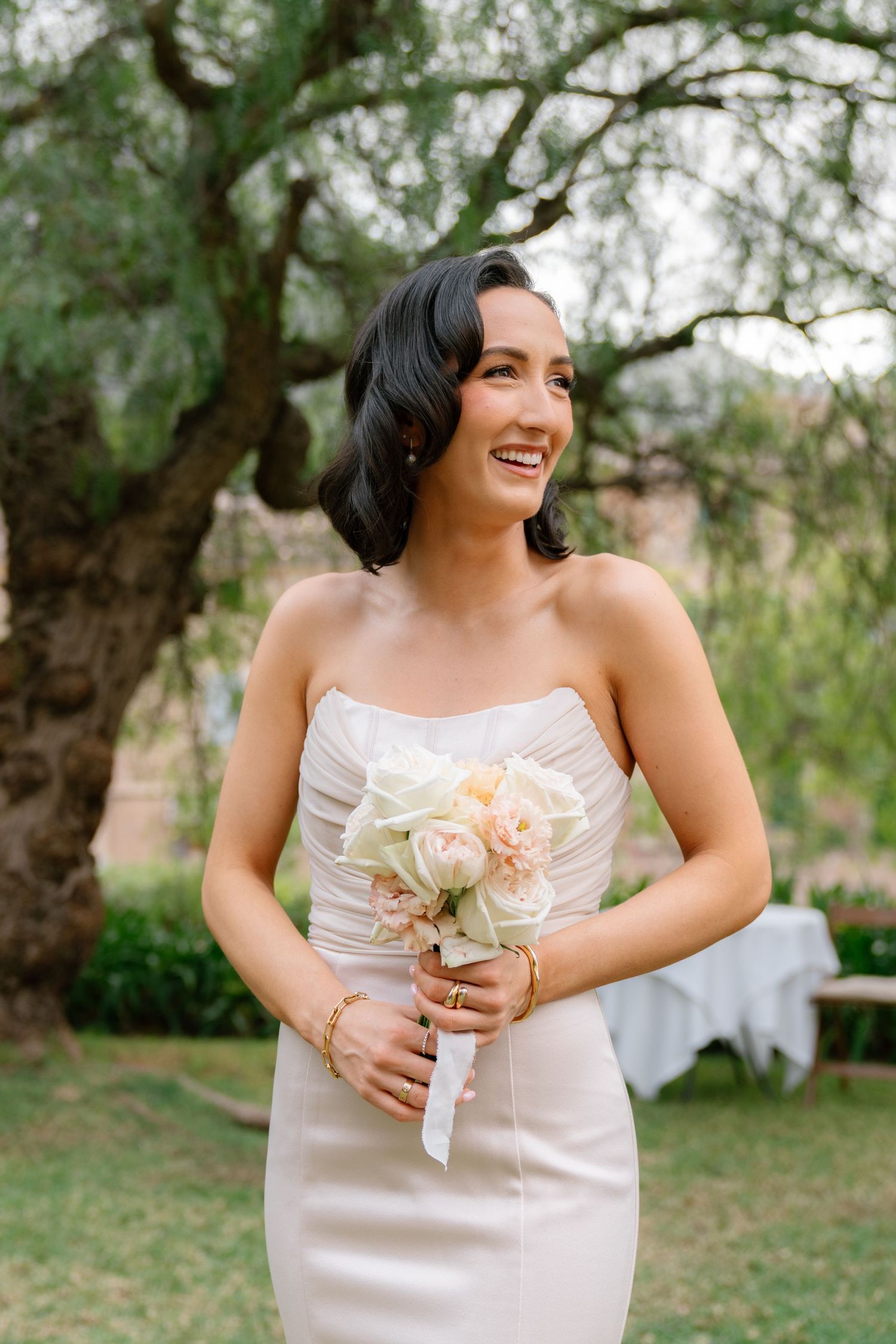 Bridesmaid in champagne dress laughs while holding delicate floral bouquet outdoors.