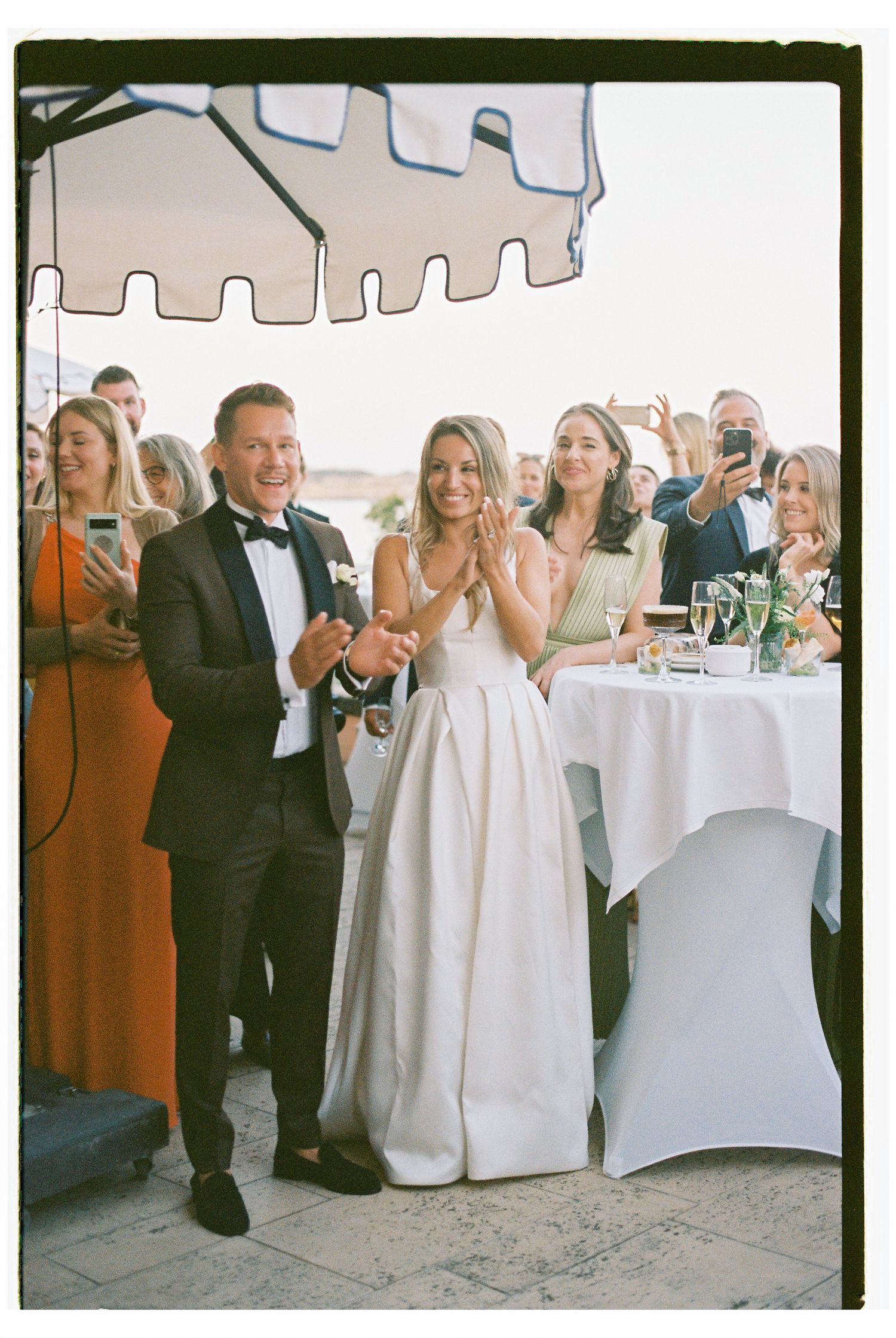 Wedding guests seated at round tables in a white tent reception space take photos with their phones.
