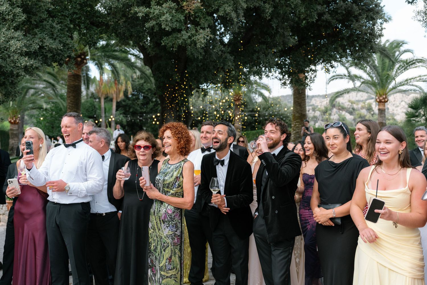 Elegantly dressed guests celebrate outdoors at a formal event, smiling and holding champagne glasses near lush palm trees.