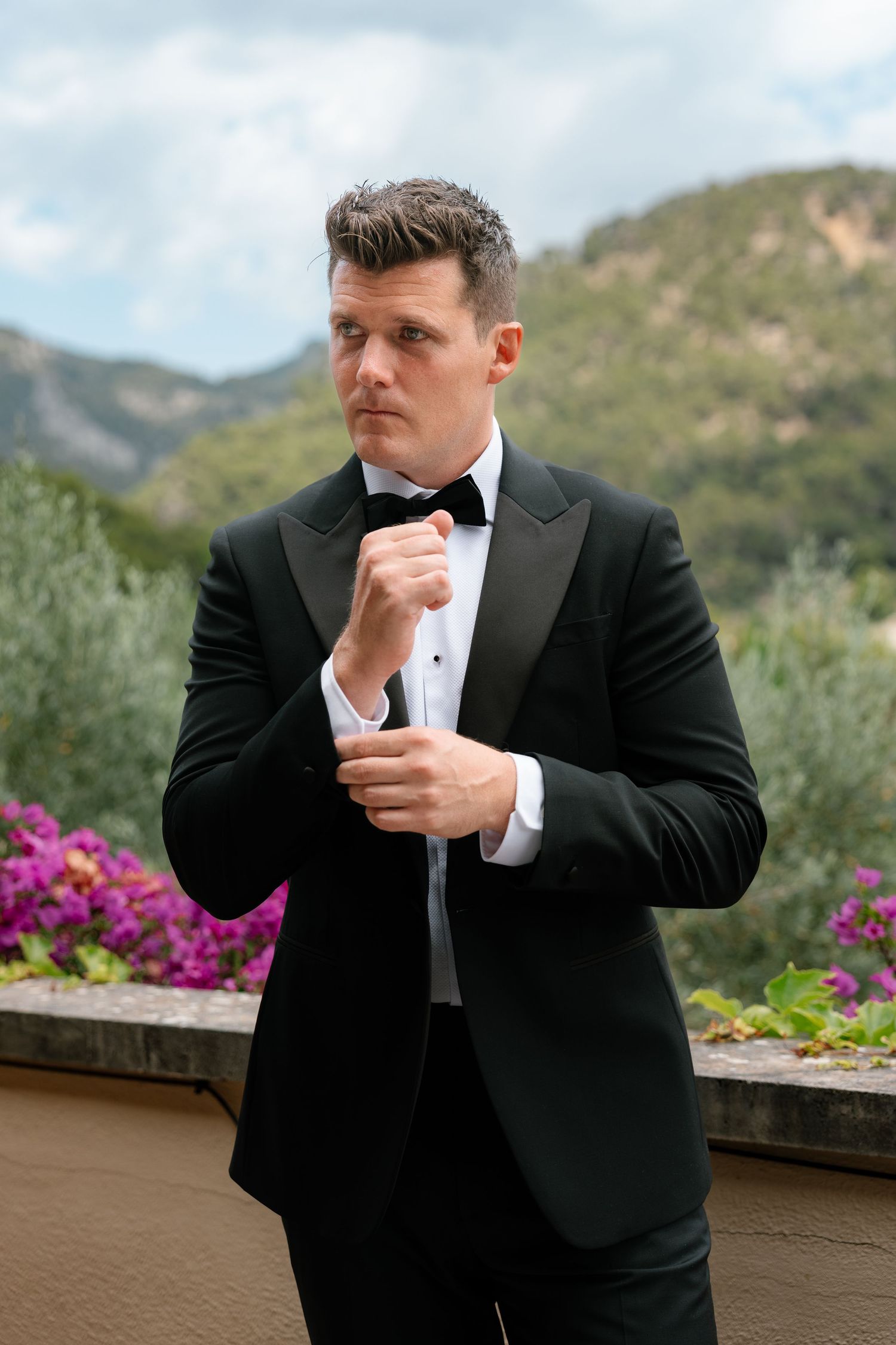 A man in a black tuxedo and bow tie adjusts his cufflinks outdoors with mountains and flowers in the background.