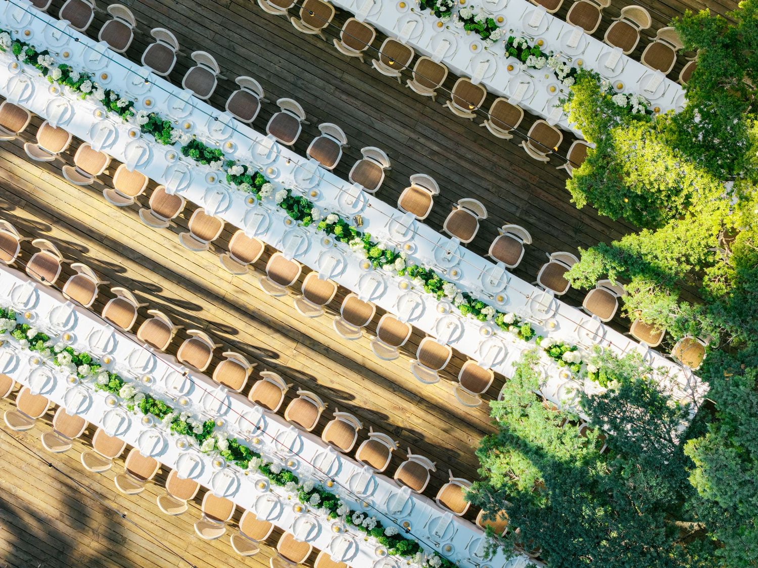 Aerial view of elegant outdoor wedding reception with long white tables, floral centerpieces, and wooden chairs on a deck.