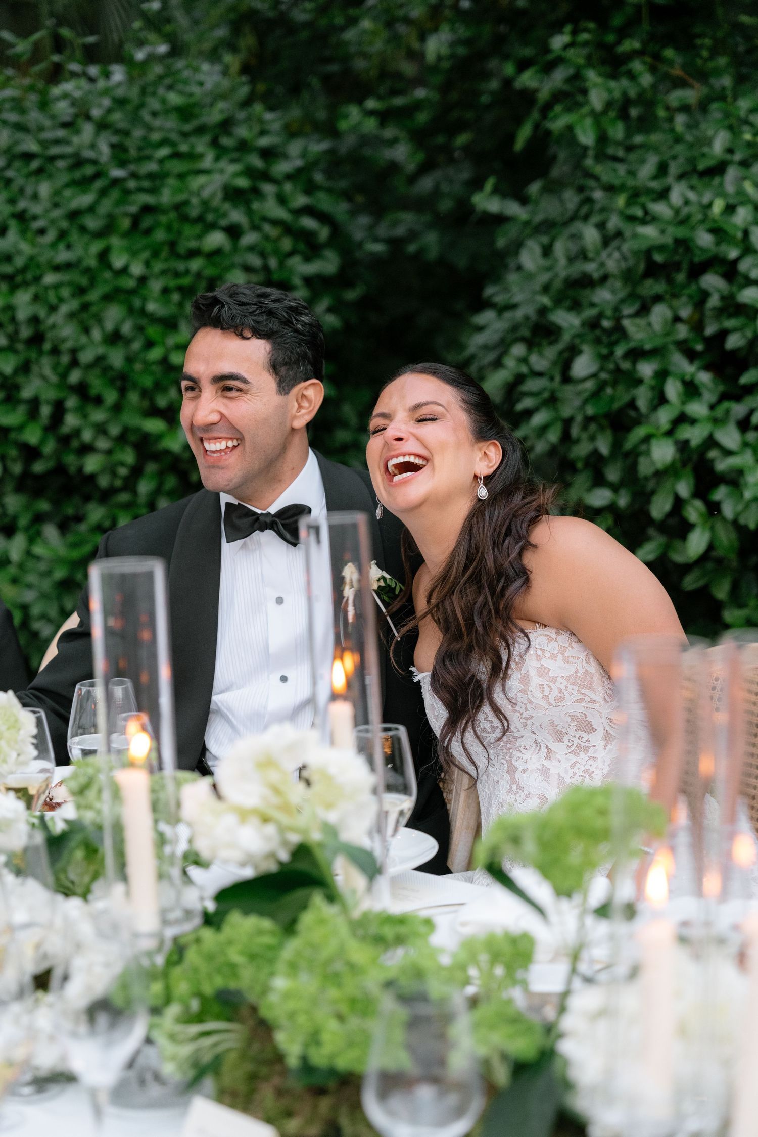 Joyful bride and groom laughing at wedding reception table with white floral centerpieces and candles outdoors.
