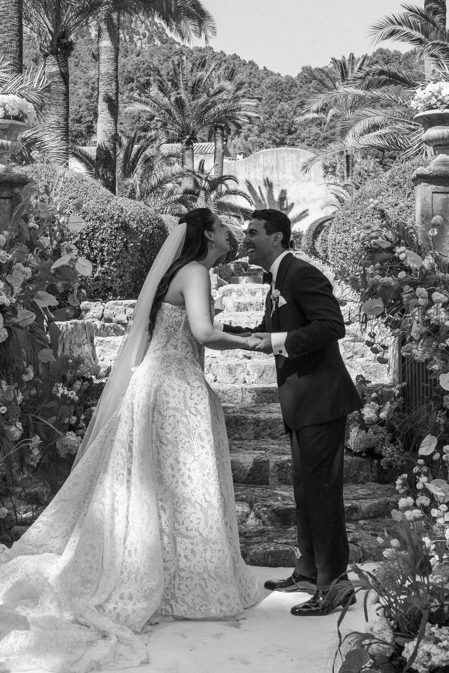 Black and white photo of a bride and groom sharing a kiss at an outdoor wedding ceremony surrounded by lush tropical gardens.
