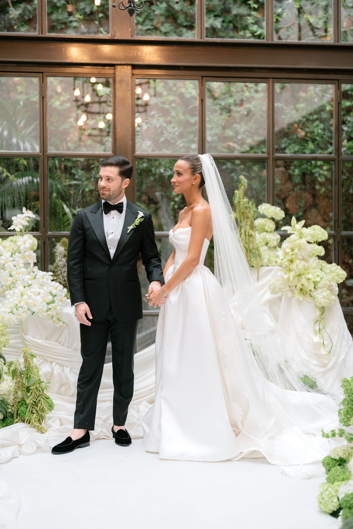 A couple in wedding attire poses for photos in a greenhouse filled with white floral arrangements.