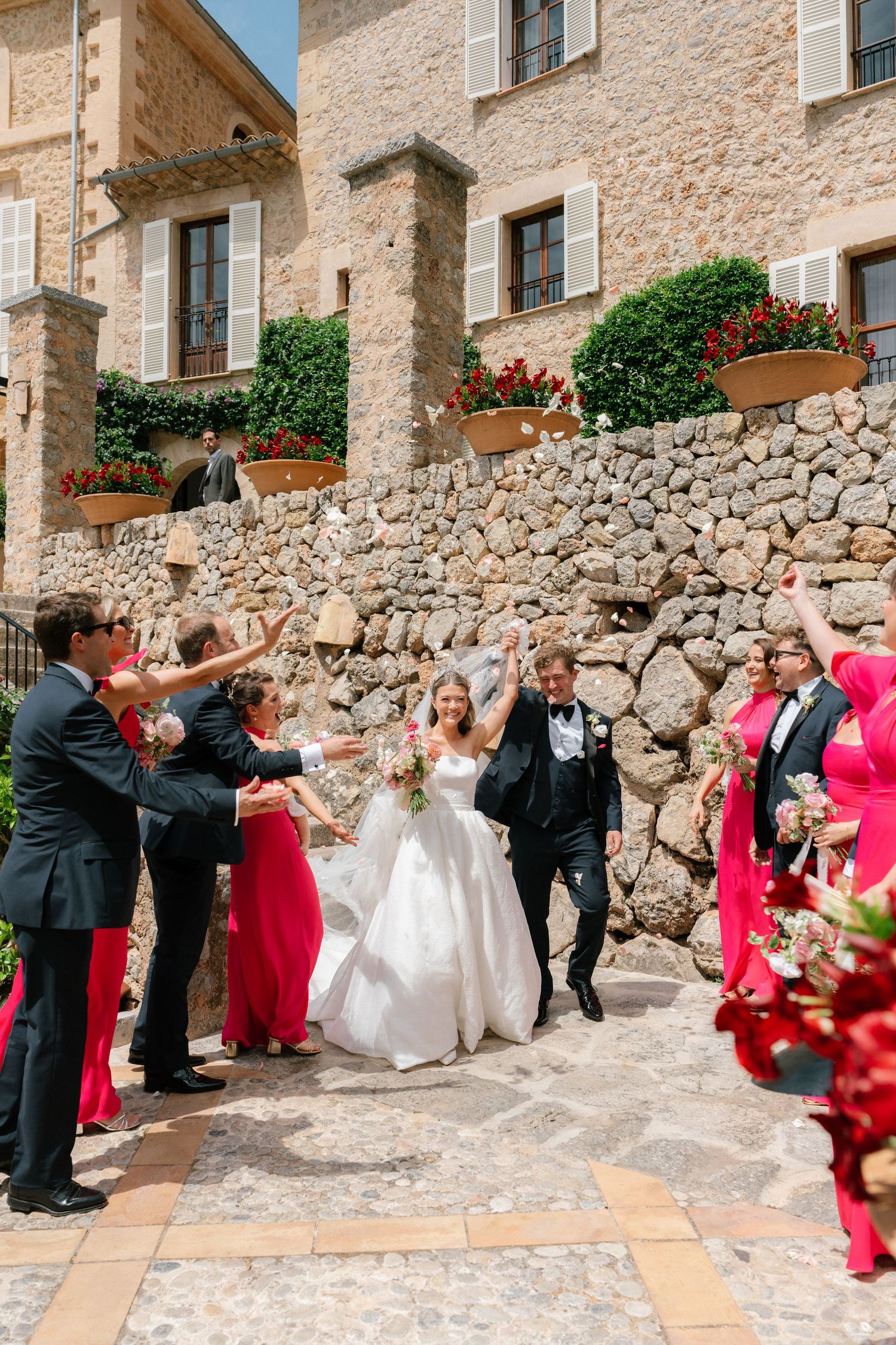 Wedding party dancing and celebrating along stone wall courtyard with red floral accents.