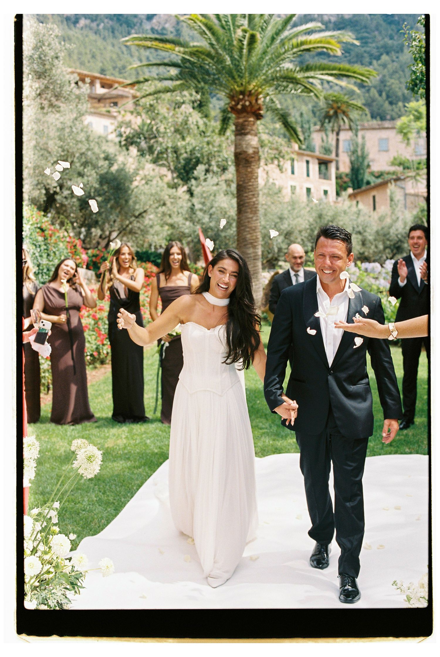 Wedding couple celebrating on a white pathway lined with palm trees as guests throw white flower petals at Belmond La Residencia, Mallorca.