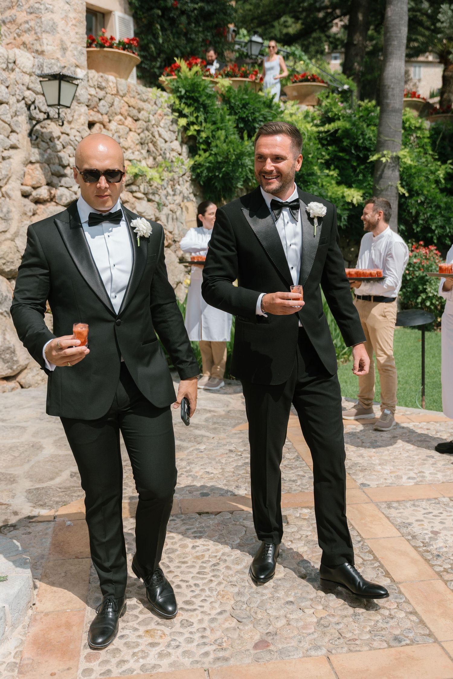 Groomsmen in black tuxedos walking along stone pathway with drinks.
