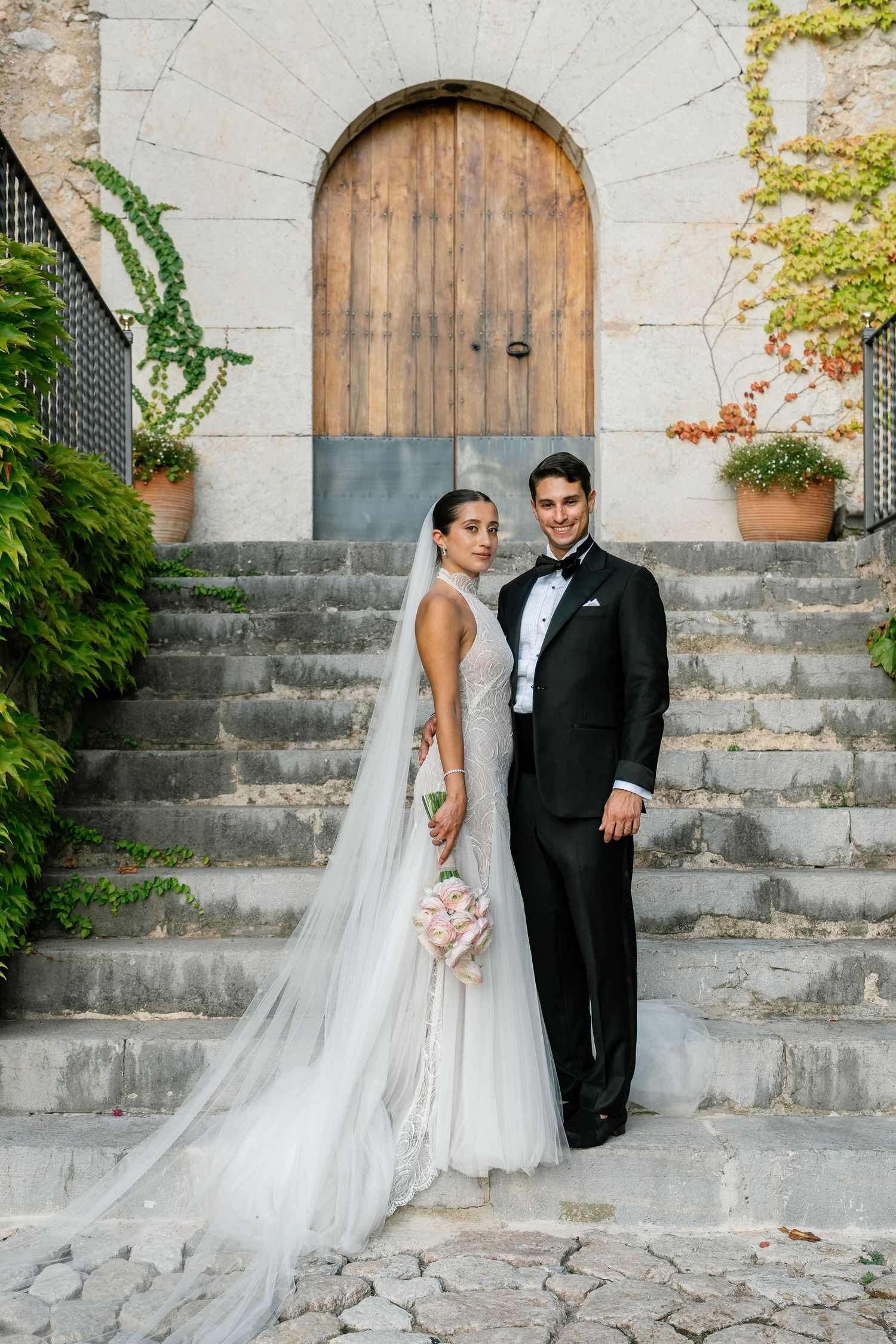 Wedding couple poses on stone steps with ivy-covered walls and a wooden door behind them.