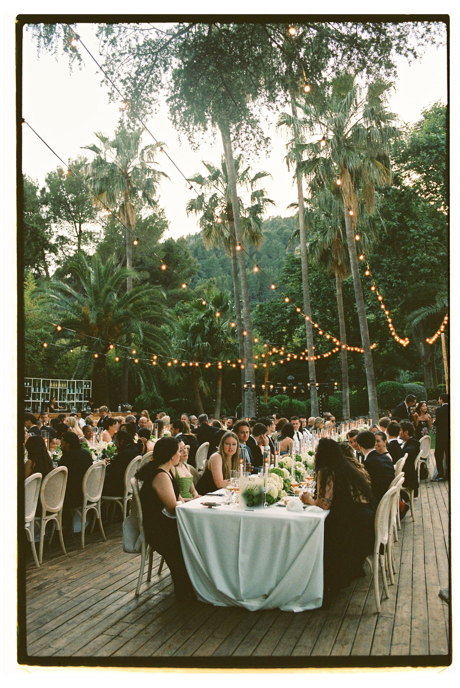 Outdoor wedding reception on wooden deck with string lights, palm trees, and guests dining at white-clothed tables at dusk.