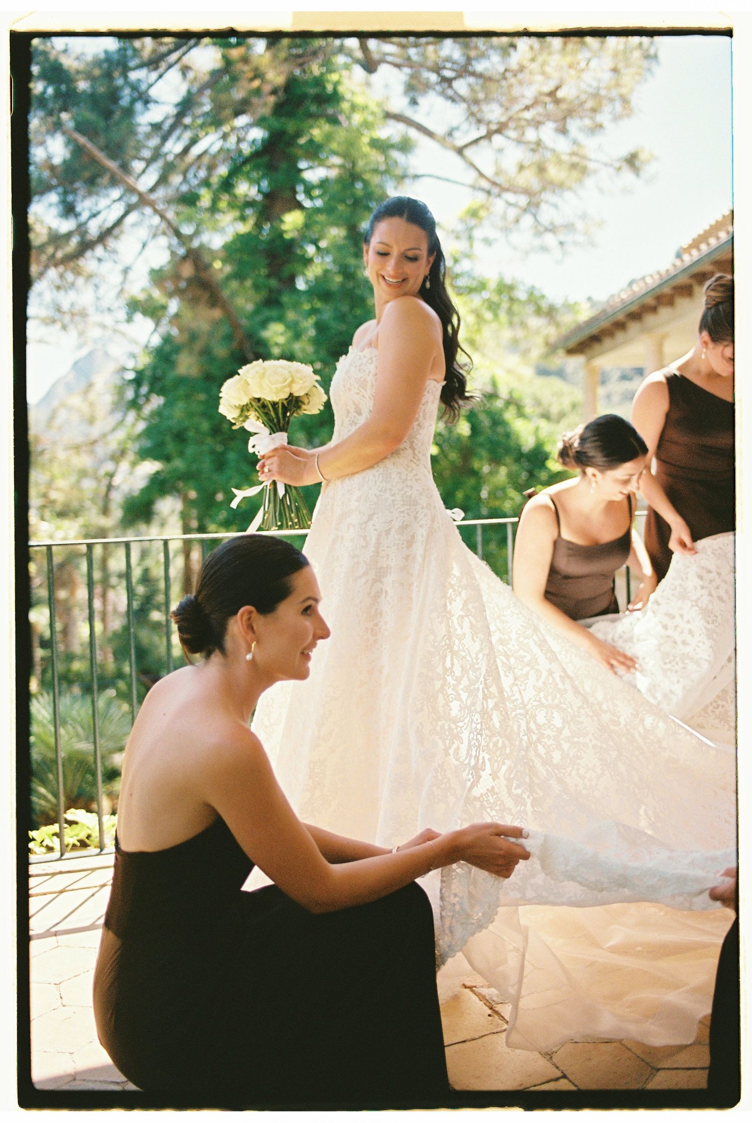 Bride in lace wedding gown holding white bouquet while bridesmaids in black dresses arrange her train outdoors.