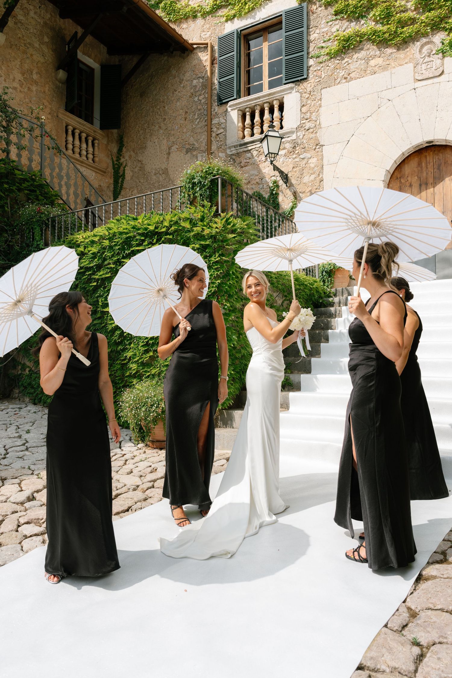 Bride in white gown with bridesmaids in black dresses holding white parasols on a white aisle at an outdoor venue.