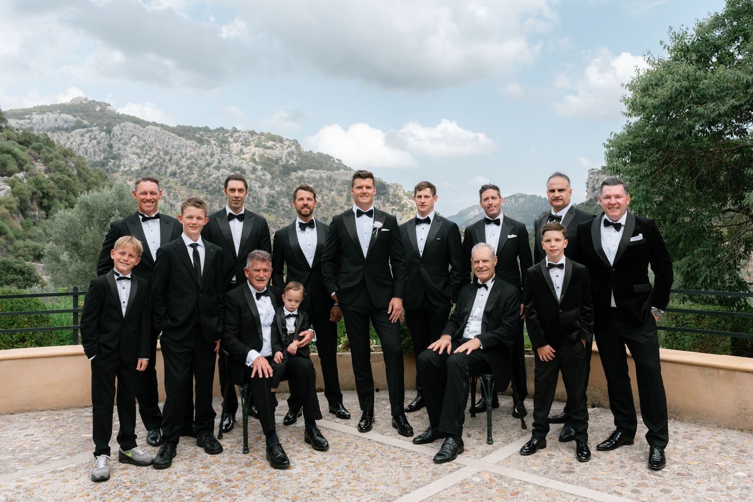 Groomsmen in black tuxedos pose together on a terrace with mountain views at a formal wedding celebration.