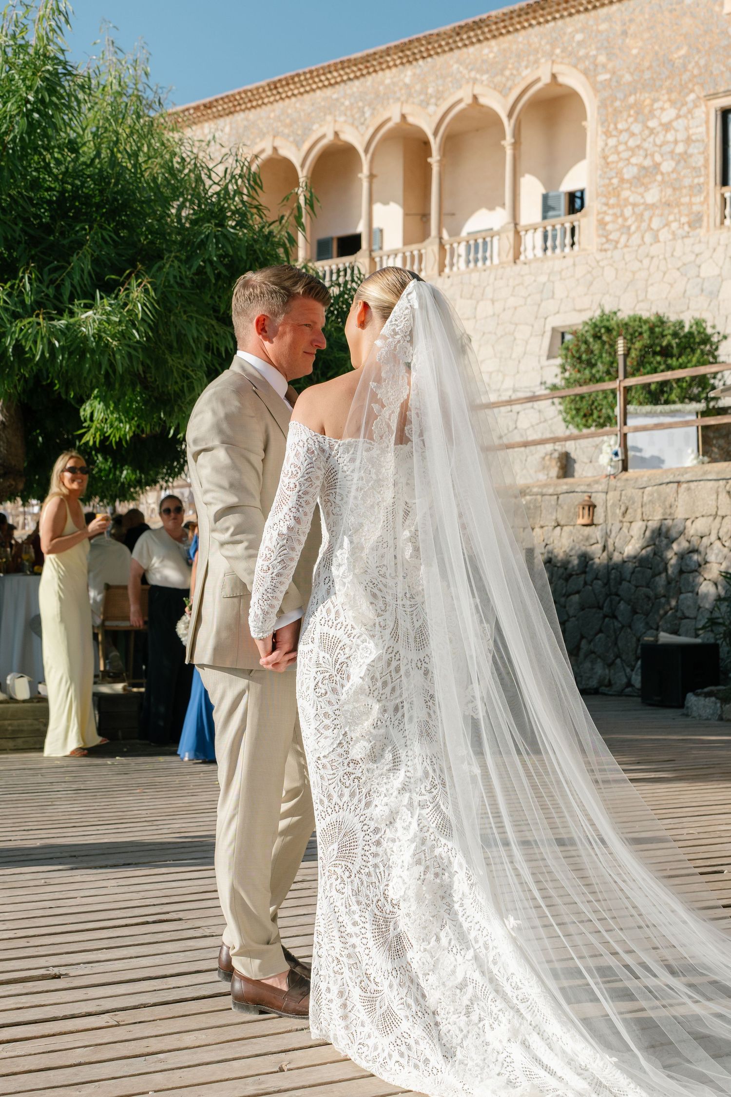 Bride in lace gown with flowing veil walks with partner at stone venue.