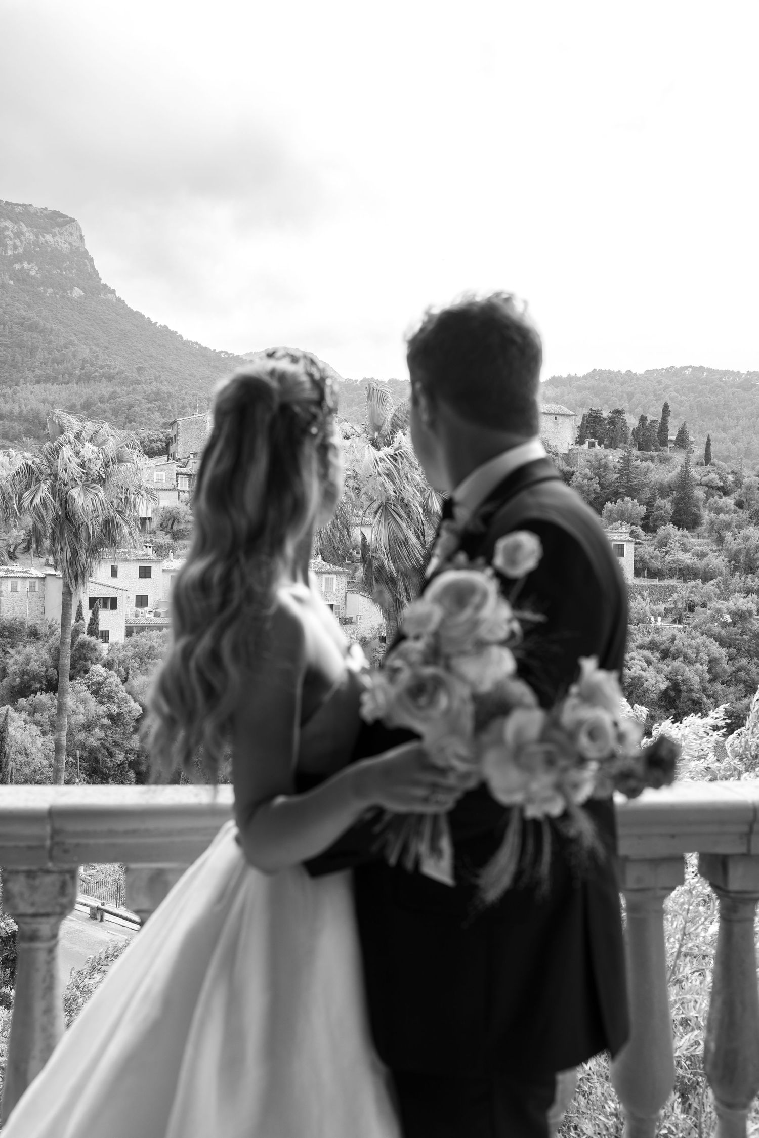 A black and white photo of newlyweds overlooking a scenic mountain village with bouquet in hand.