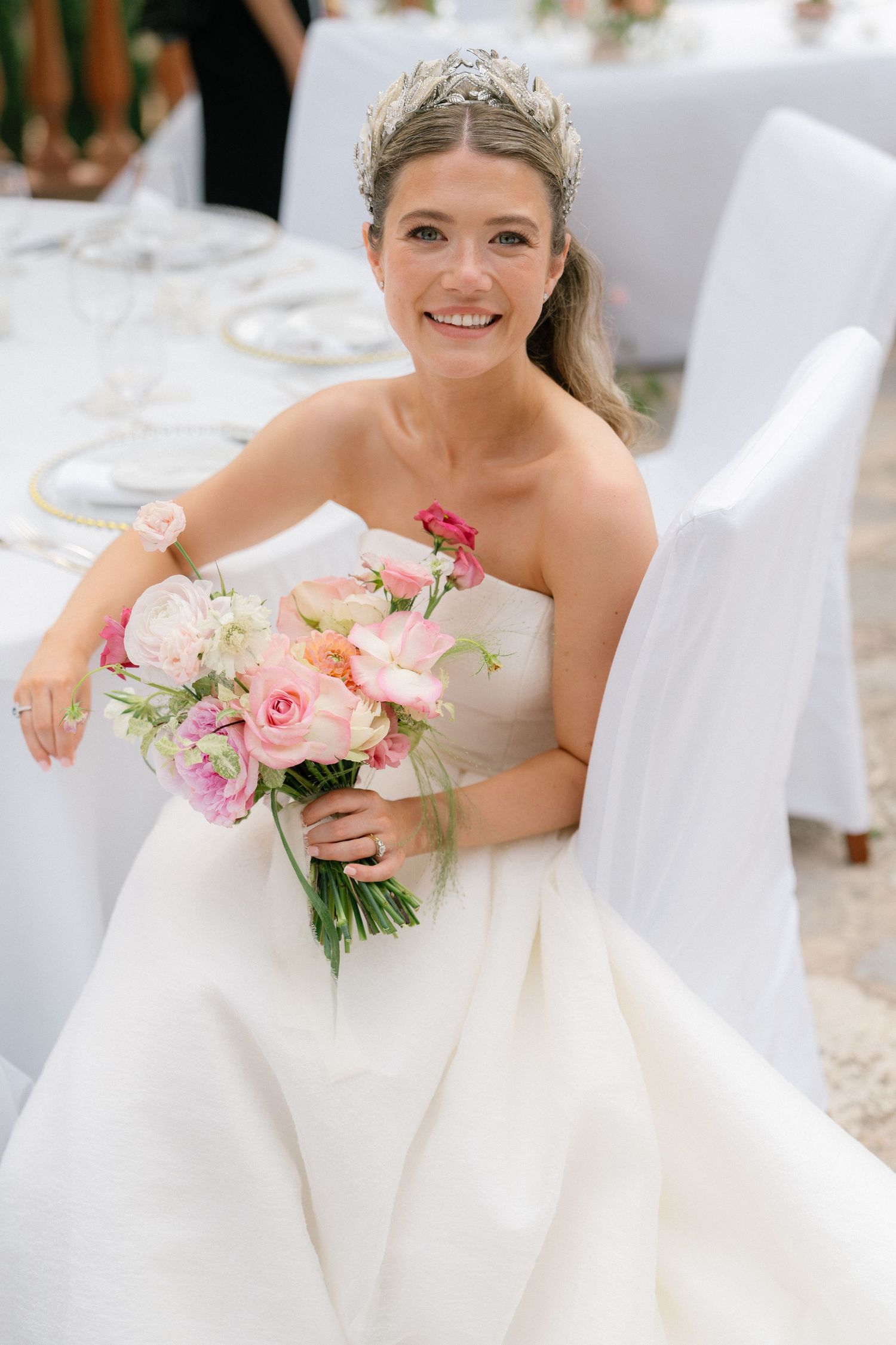 Elegant bride in white gown poses with pink roses against white decor.