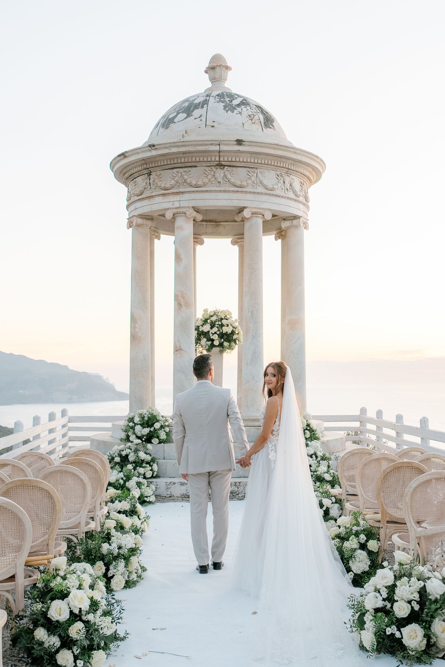 Romantic seaside wedding ceremony setup with white gazebos and floral arrangements overlooking the ocean at sunset.