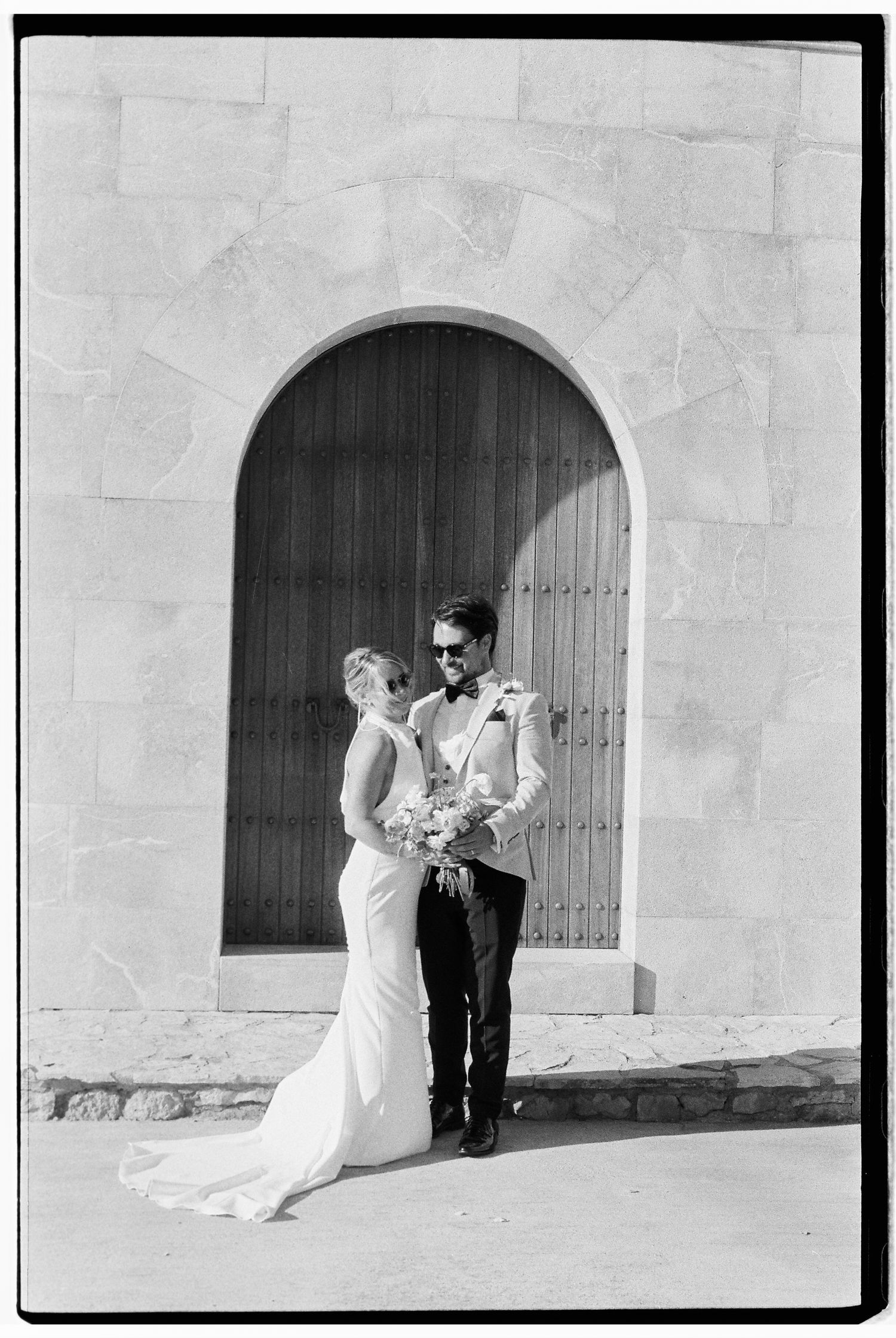 Wedding couple posing against stone wall with arched doorway in black and white.