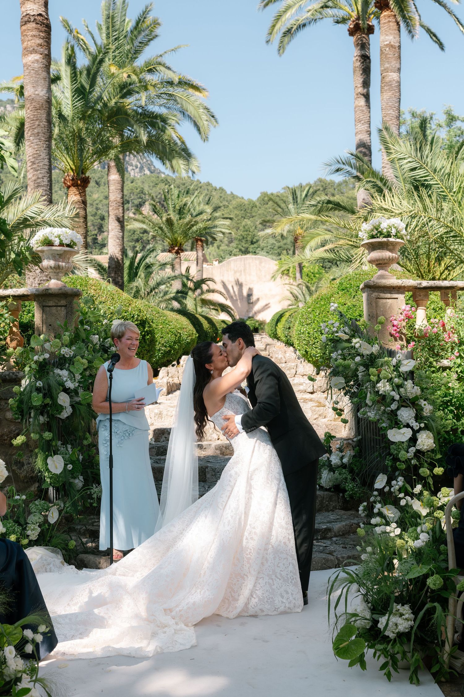 Bride and groom share first kiss at outdoor wedding ceremony surrounded by palm trees and white floral arrangements.