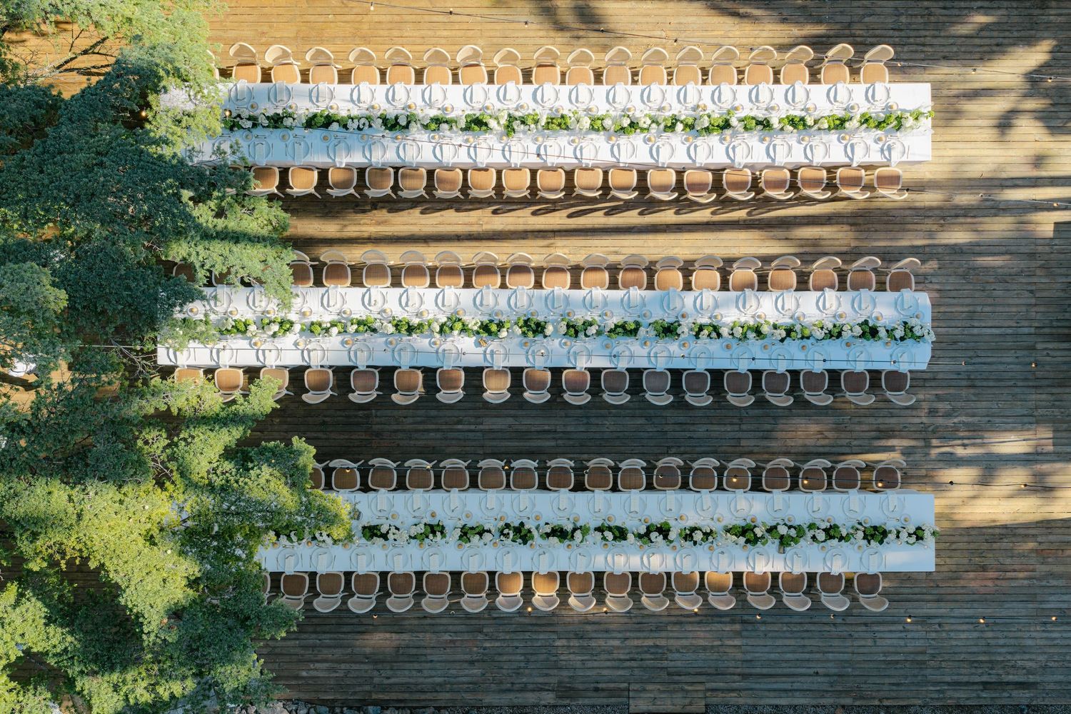 Aerial view of three long outdoor dining tables with white linens and wooden chairs on a wooden deck.