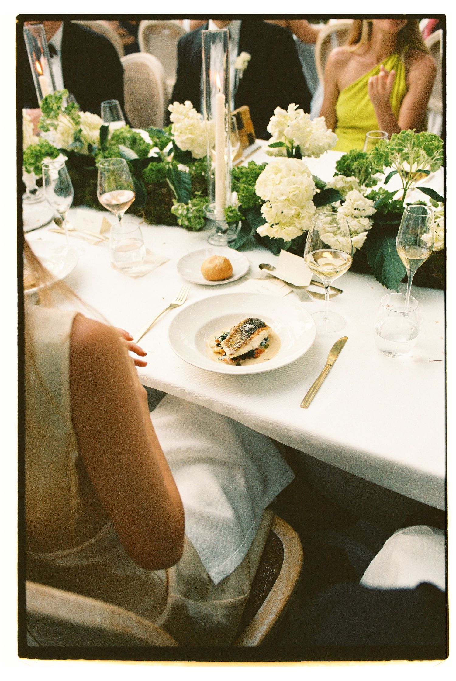Elegant wedding reception table with white floral centerpieces, wine glasses, and plated fish dish on white linen.