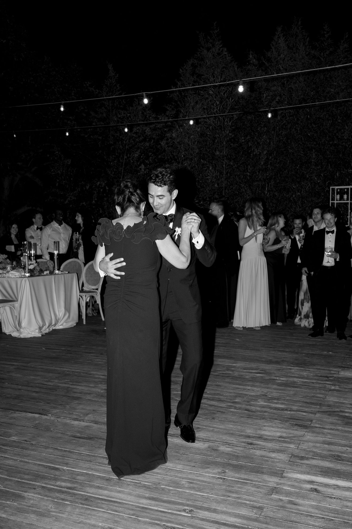 Black and white photo of a couple sharing a romantic dance at an outdoor wedding reception under string lights.