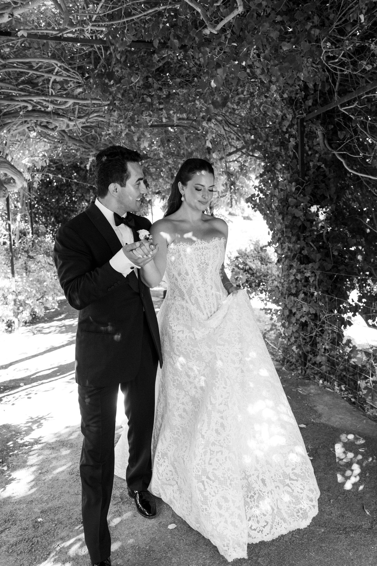 Black and white wedding photo of bride in lace gown and groom in tuxedo walking under ivy archway.