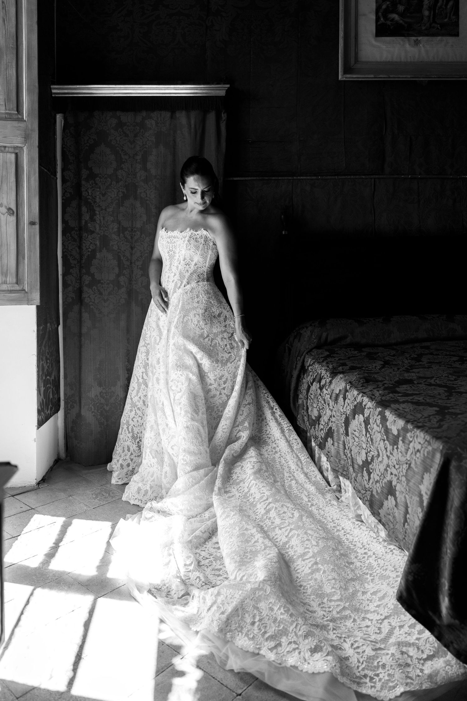 Bride in strapless lace wedding gown standing in sunlit room, black and white portrait photography.