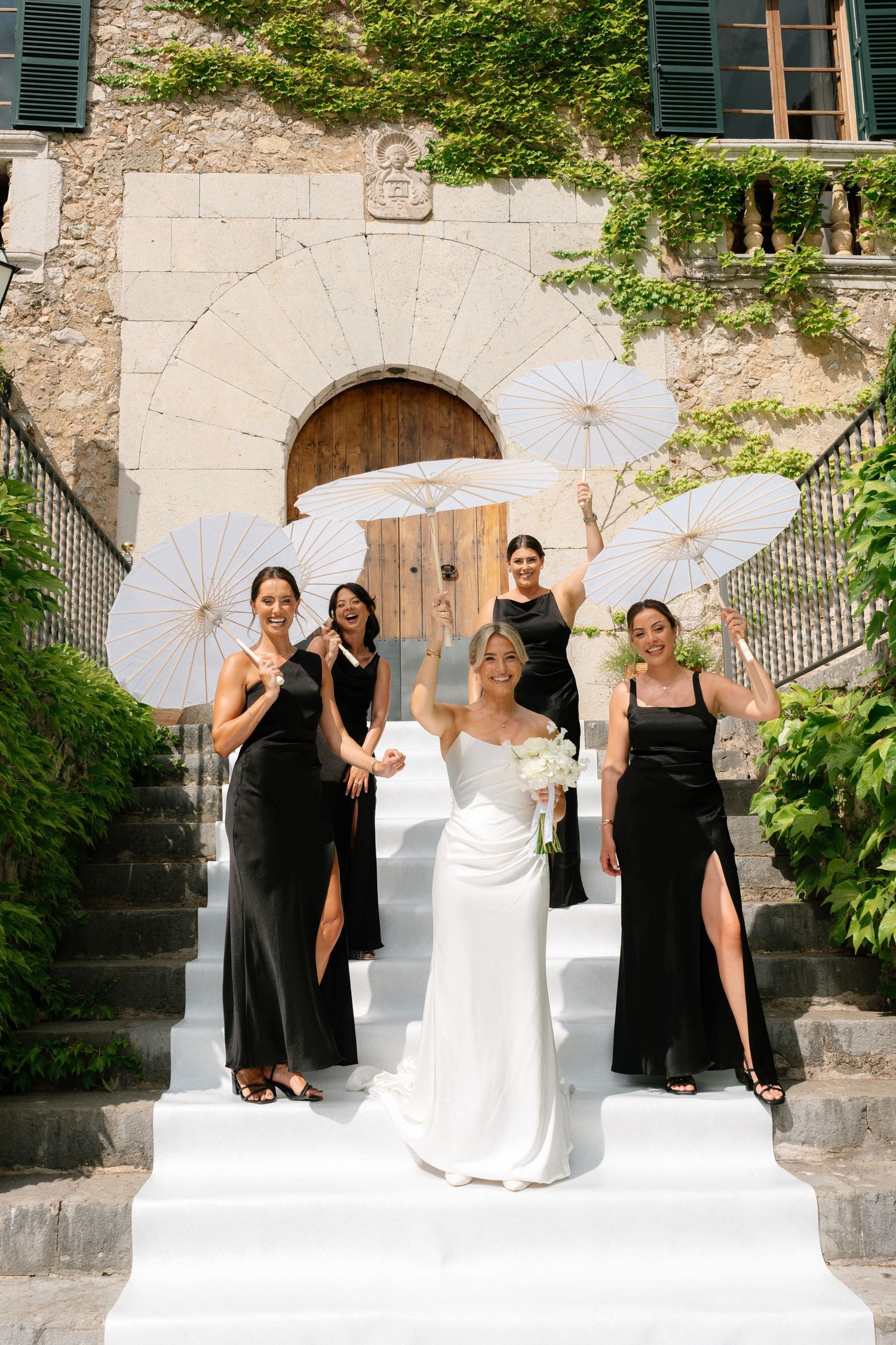 Bride in white gown with bridesmaids in black dresses holding white parasols on stone steps of elegant venue.