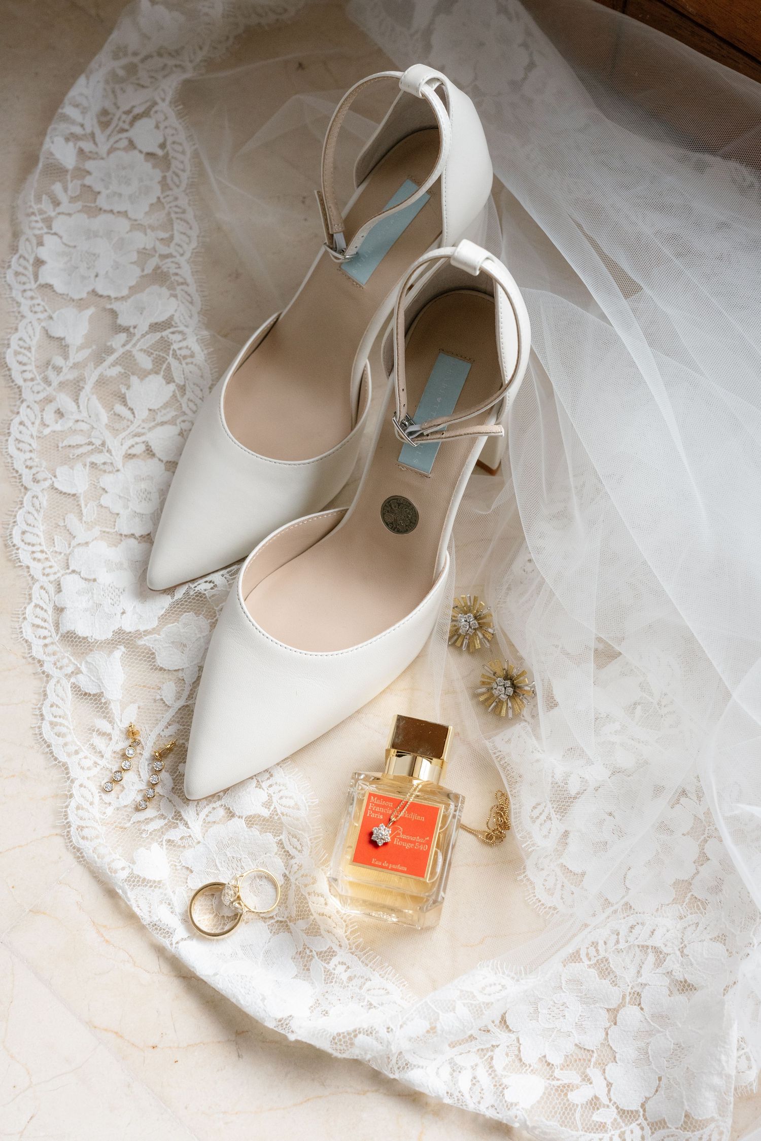 White bridal heels with ankle straps rest on lace veil beside gold earrings and perfume bottle.