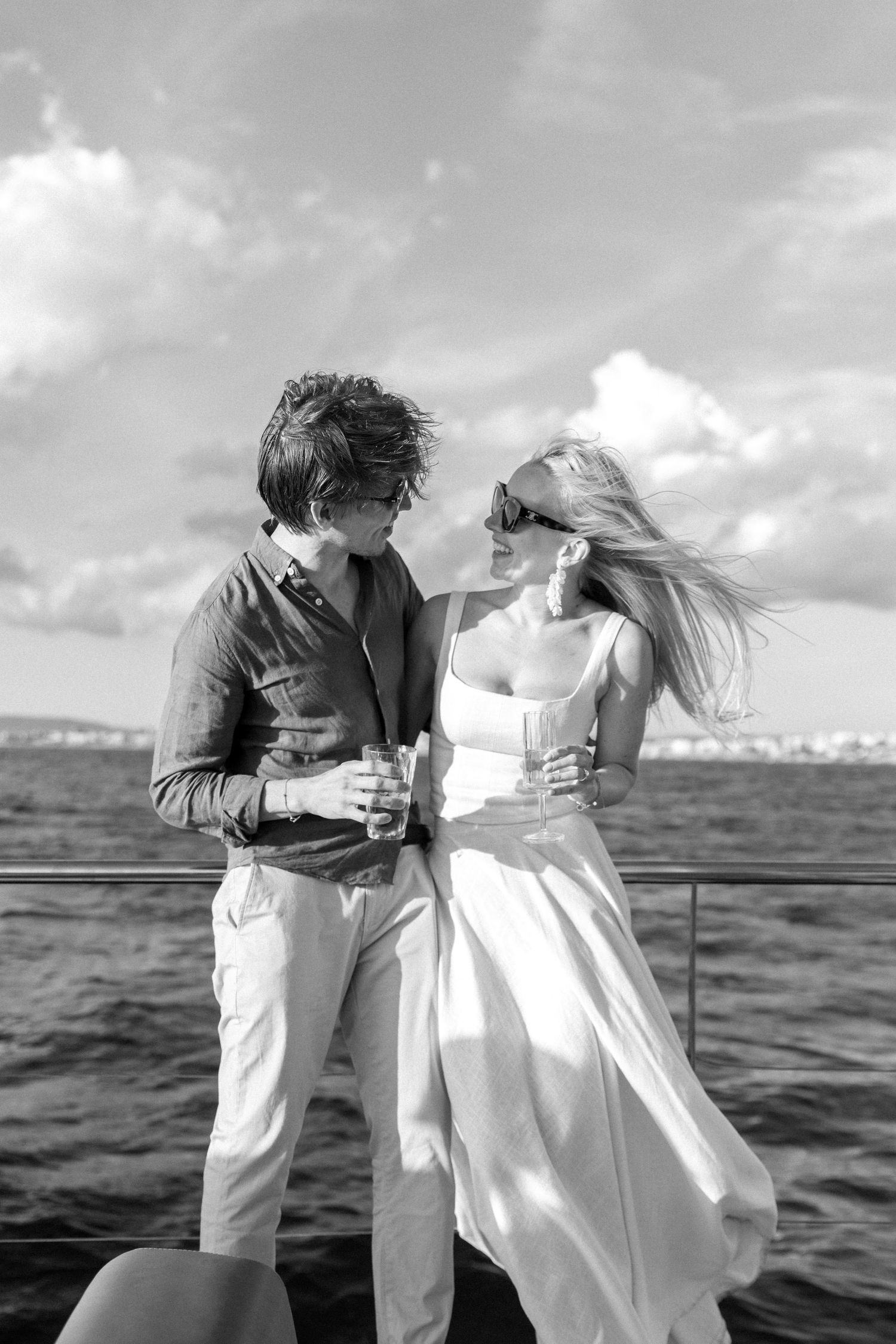 A couple in white and beige clothing embraces while dancing on a boat with the sea in the background.