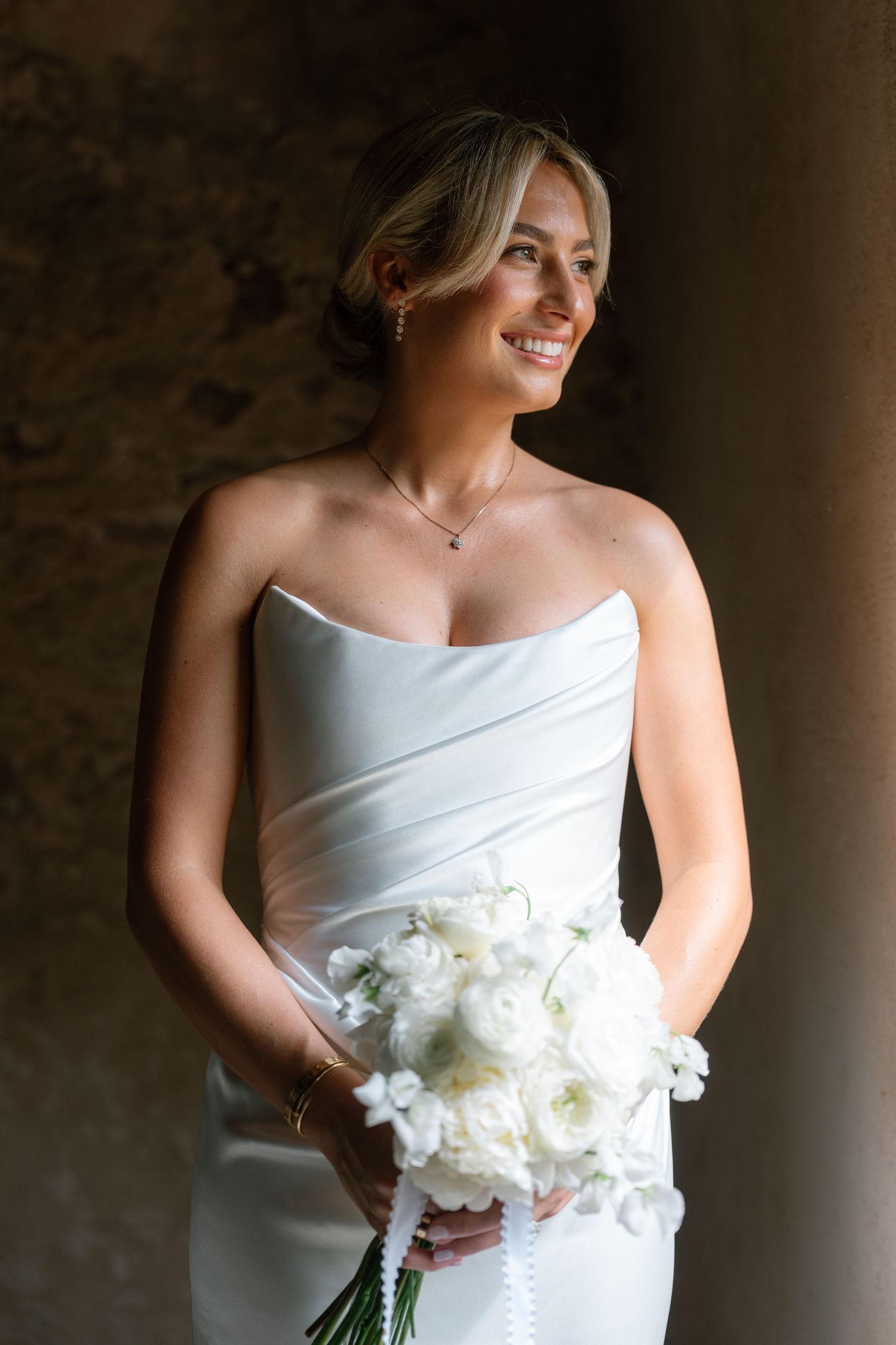 Smiling bride in strapless white gown holds white bouquet of roses, standing in soft light against stone wall.