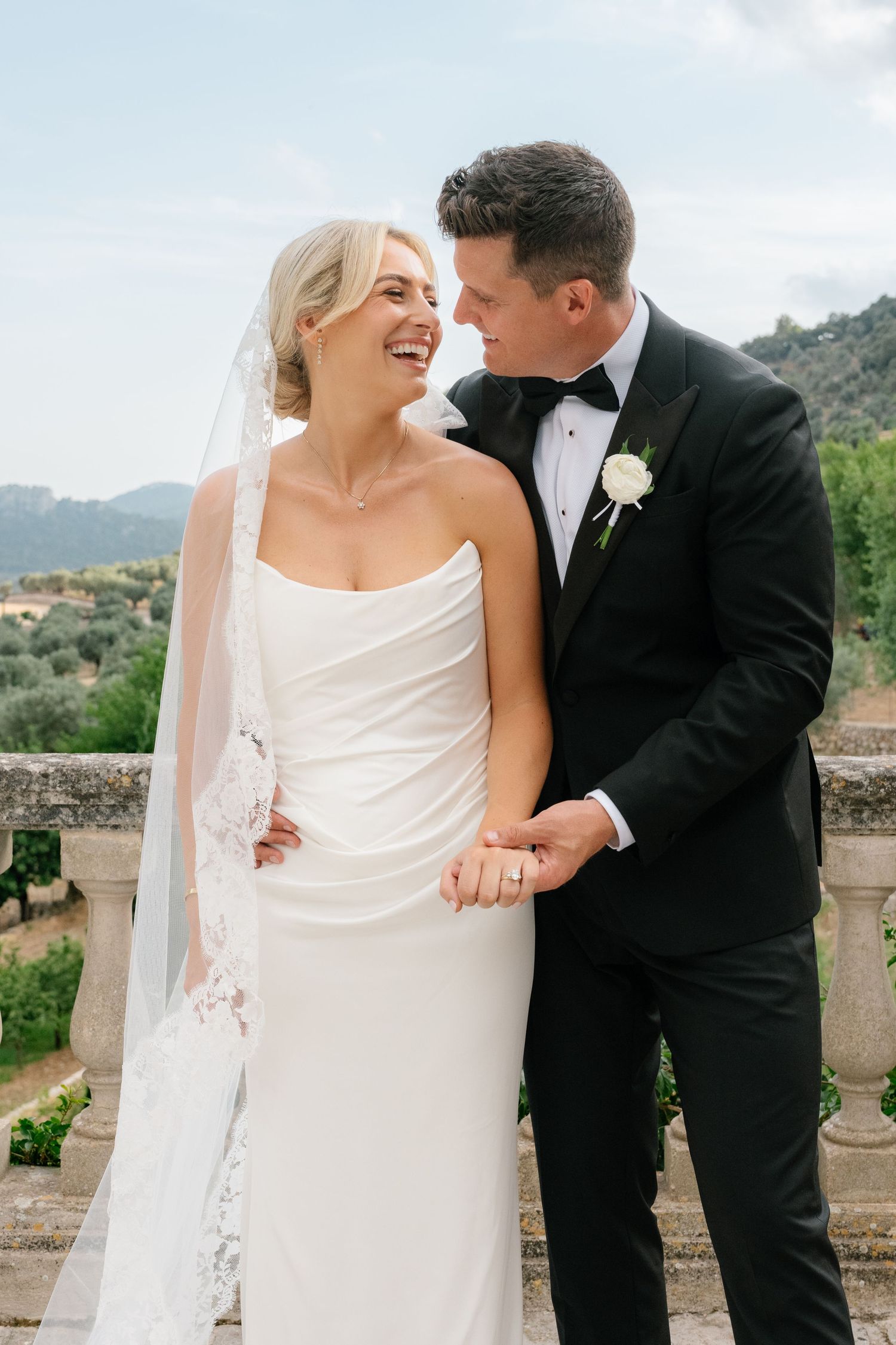 Bride in white gown and veil laughs with groom in black tuxedo outdoors with mountain views.