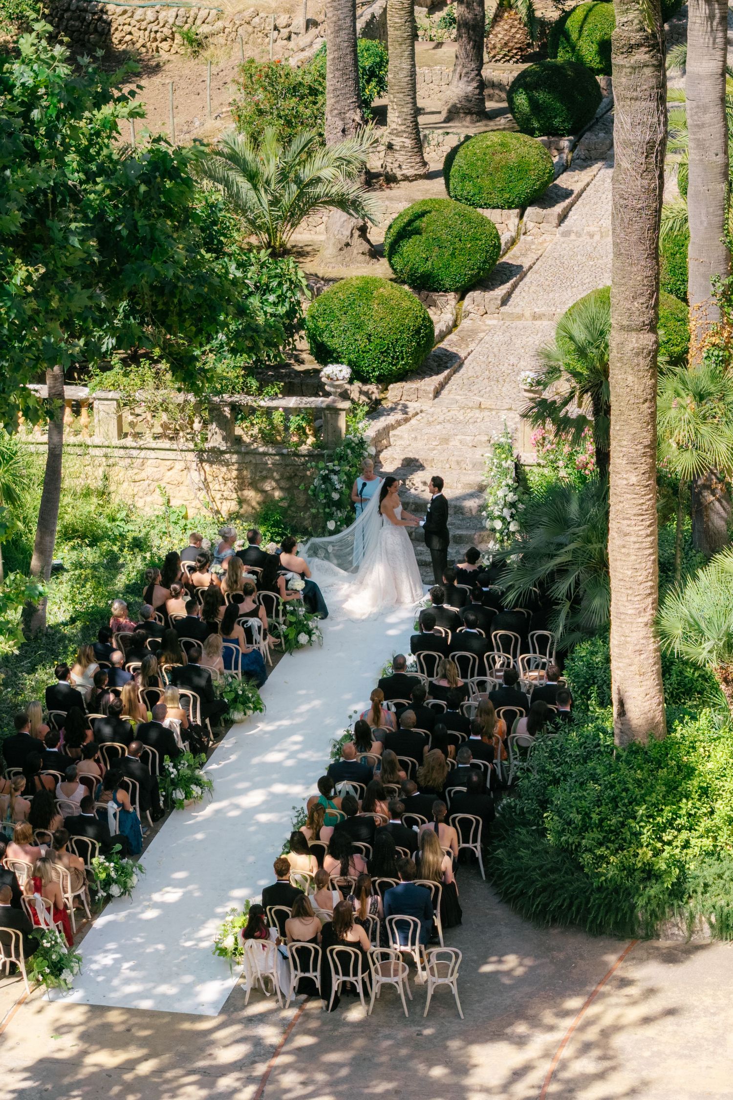 Aerial view of an outdoor wedding ceremony with guests seated along a white aisle in a lush garden setting.