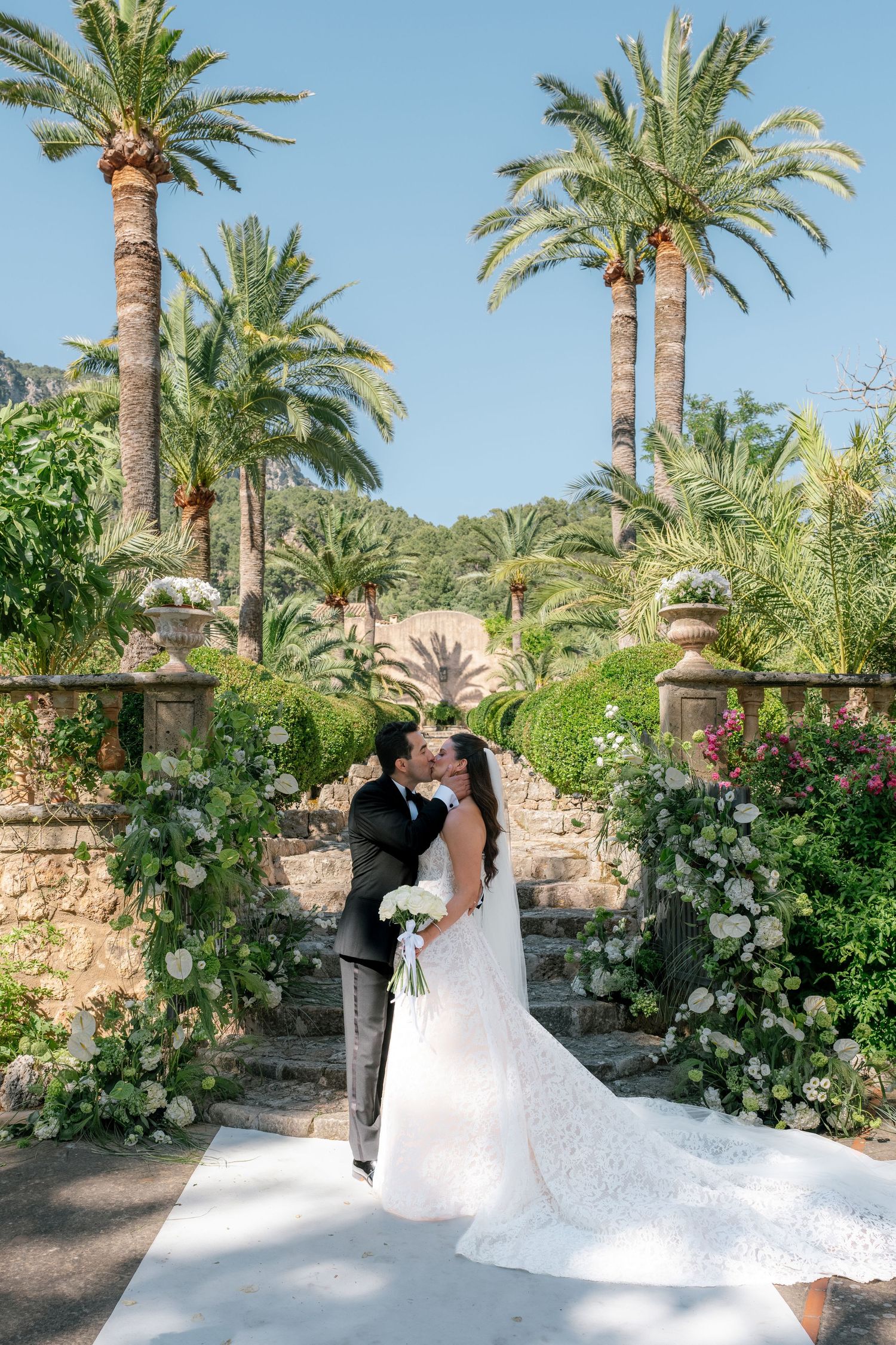 Bride and groom kissing at outdoor wedding ceremony surrounded by lush palm trees and elegant floral arch.