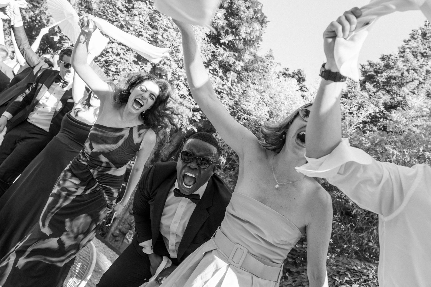 Black and white photo of joyful wedding party celebrating outdoors, bride in gown cheering with guests in formal attire.