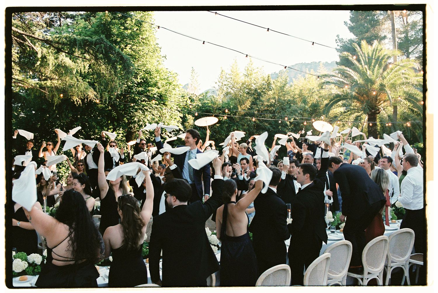Outdoor wedding reception guests celebrate raising white napkins in a lush garden venue with string lights.