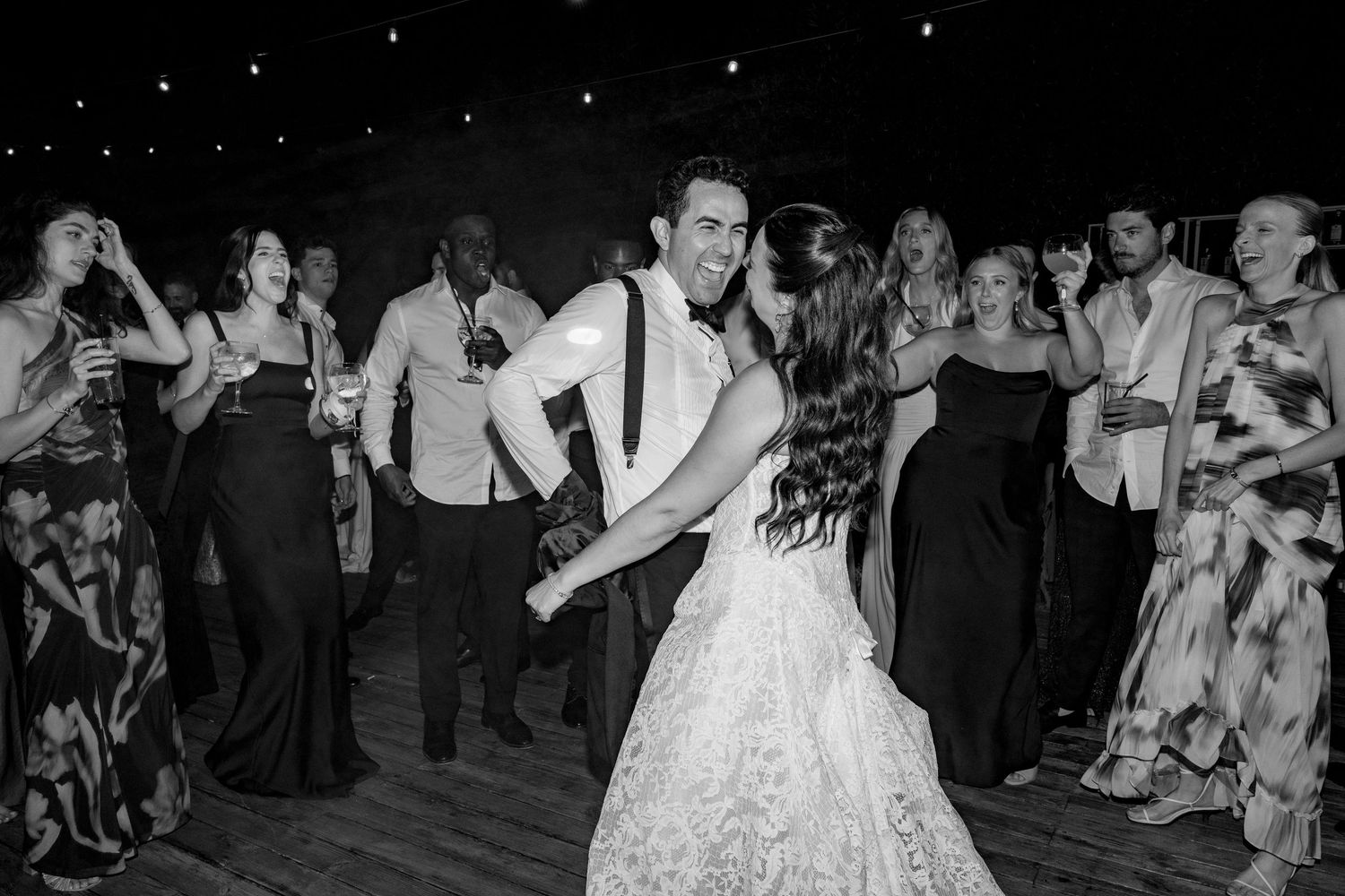 Black and white photo of a bride and groom sharing their first dance surrounded by guests at a wedding reception.
