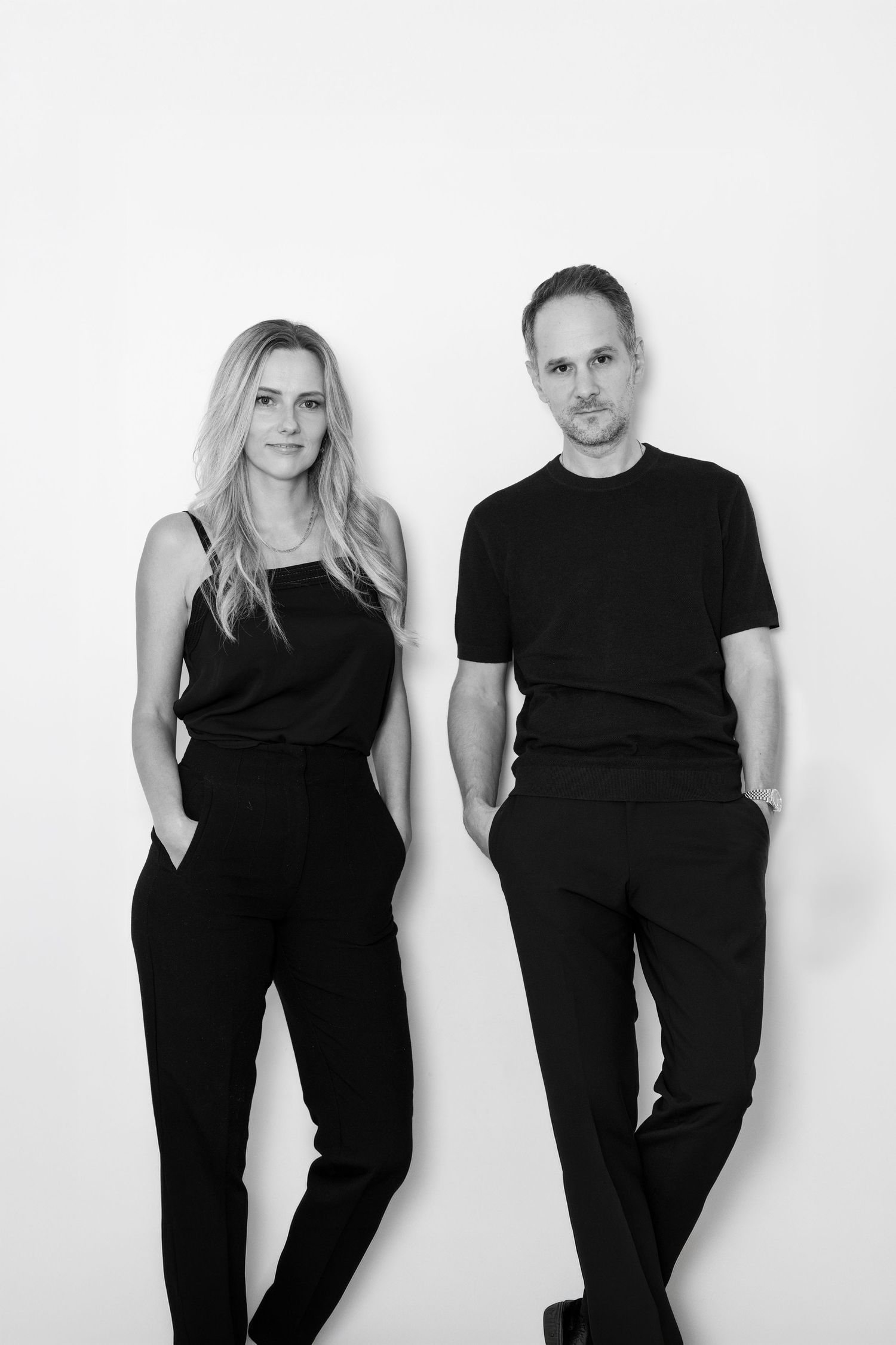 Black and white portrait of a woman and a man in matching black outfits standing casually against a plain white wall.