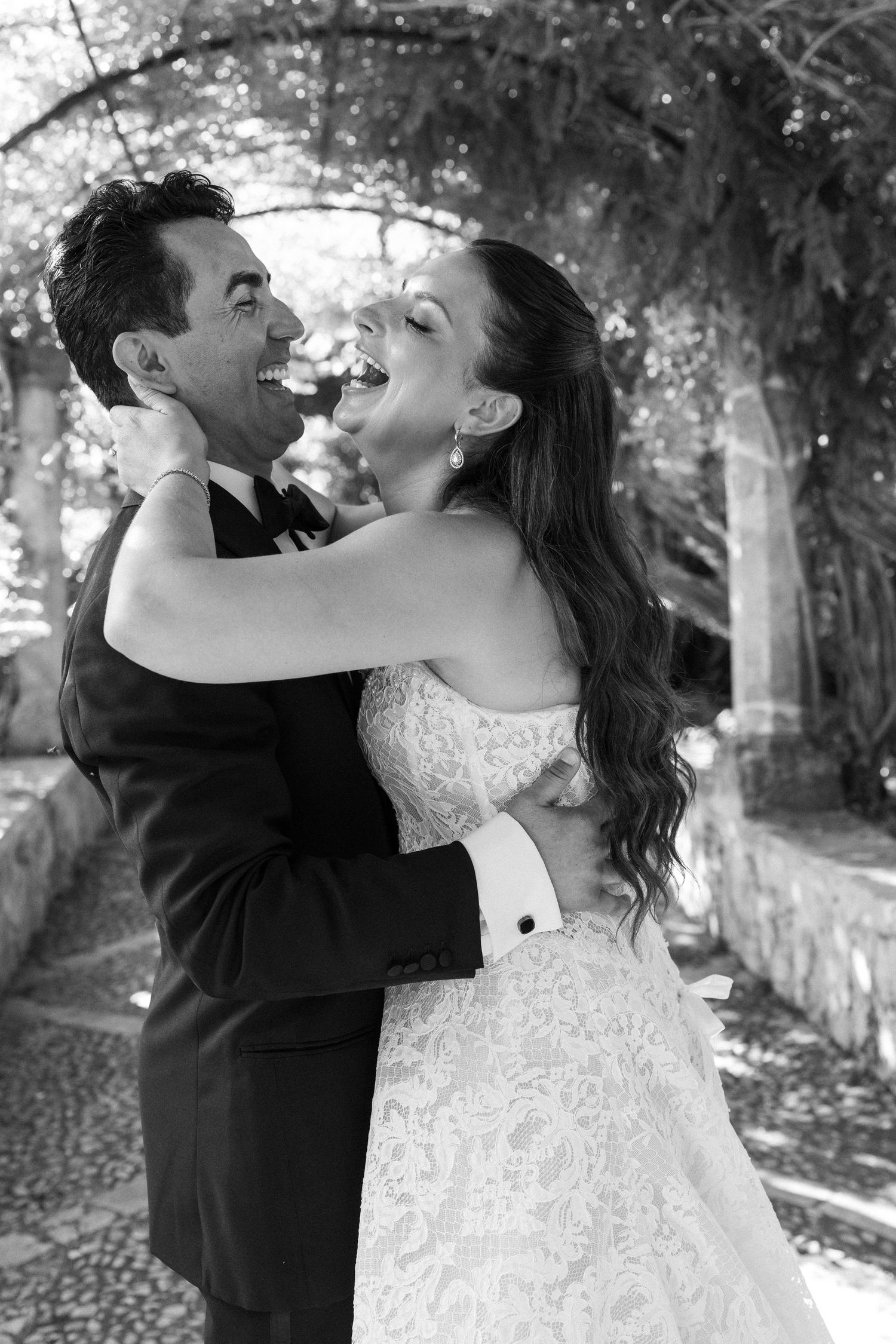 Black and white photo of a joyful bride and groom embracing and laughing together under a vine-covered archway.
