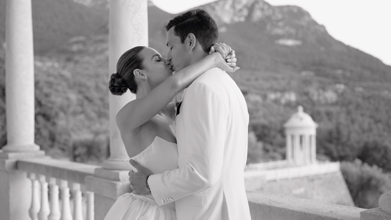 Black and white photo of a couple sharing a romantic kiss on a balcony with mountain scenery at their wedding.