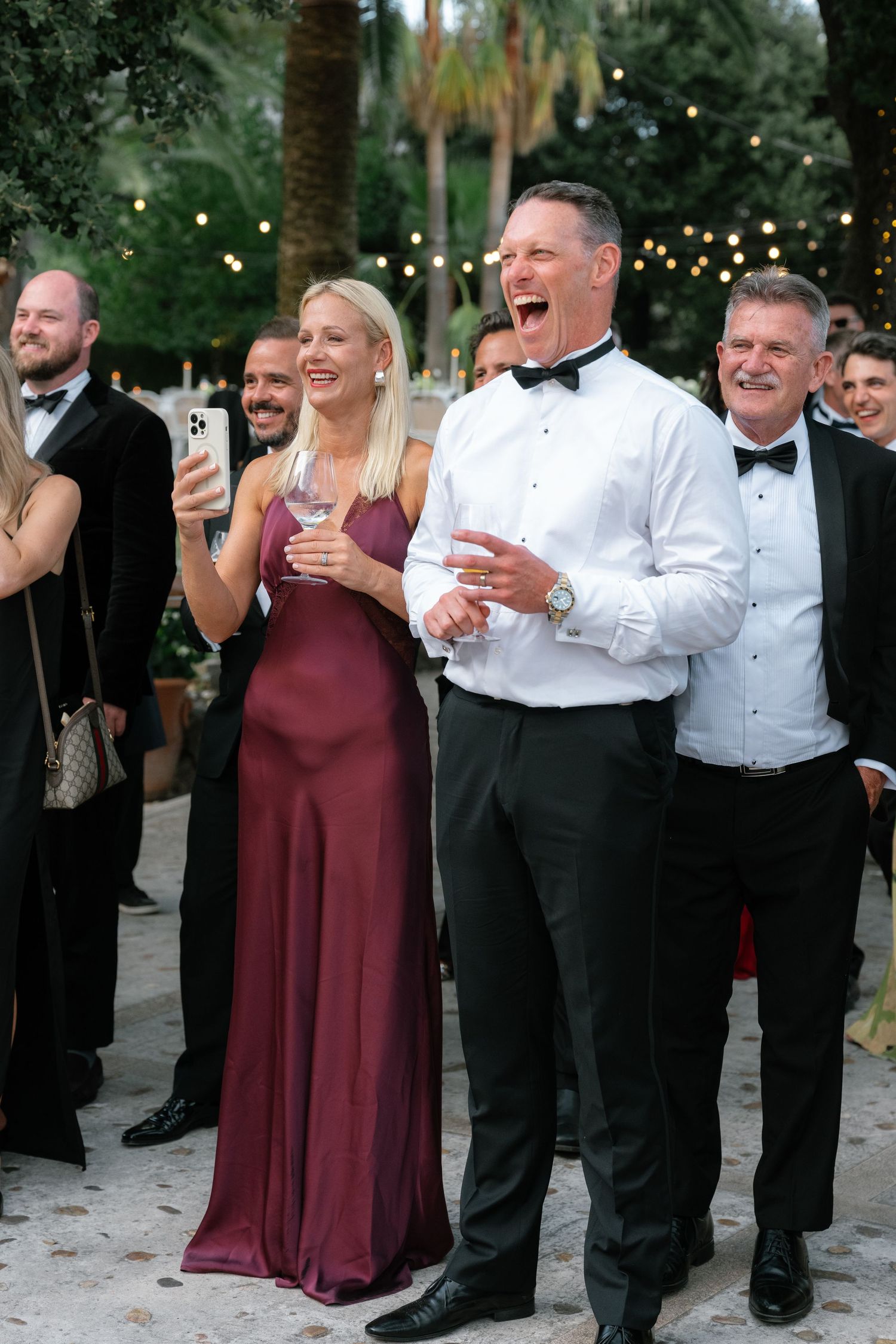 Guests laughing and celebrating at an elegant outdoor evening event, dressed in formal black tie attire.