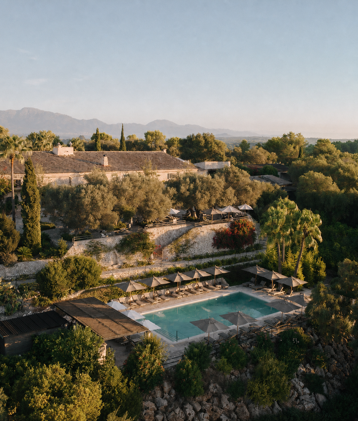 Aerial view of a luxury Mediterranean resort Finca Serena with pool, sun loungers, and mountain backdrop surrounded by lush greenery.
